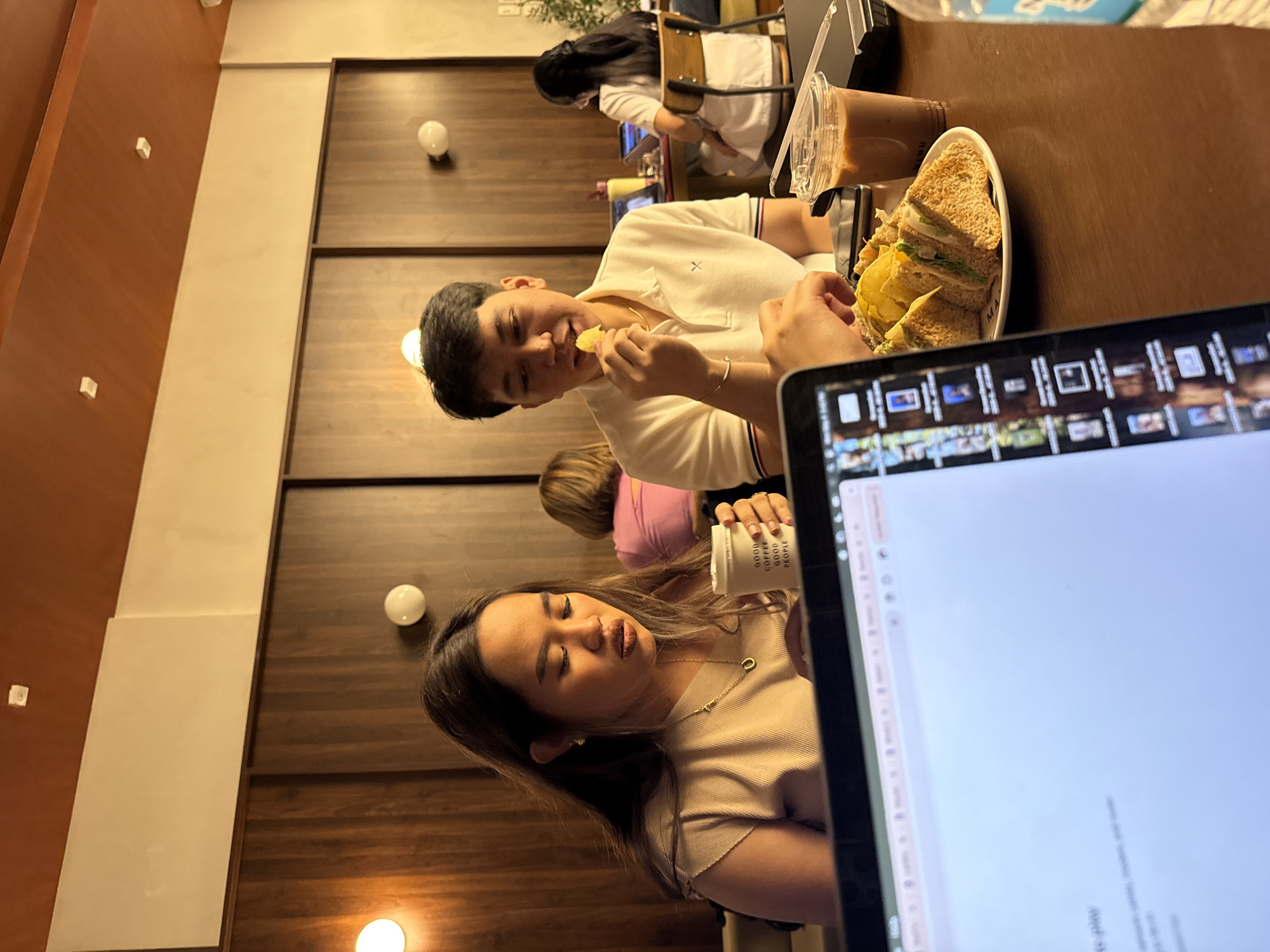 The image depicts a group of people seated at a table in an indoor setting, possibly a café. A man is eating a chip, and a woman is holding a cup labeled "GOOD COFFEE GOOD PEOPLE". A plate of sandwiches and a drink are on the table. A laptop is in the foreground. Other people are present in the background. The café features a brown and beige color scheme with wooden wall panels and spherical light fixtures. The location is Davao City, Philippines.