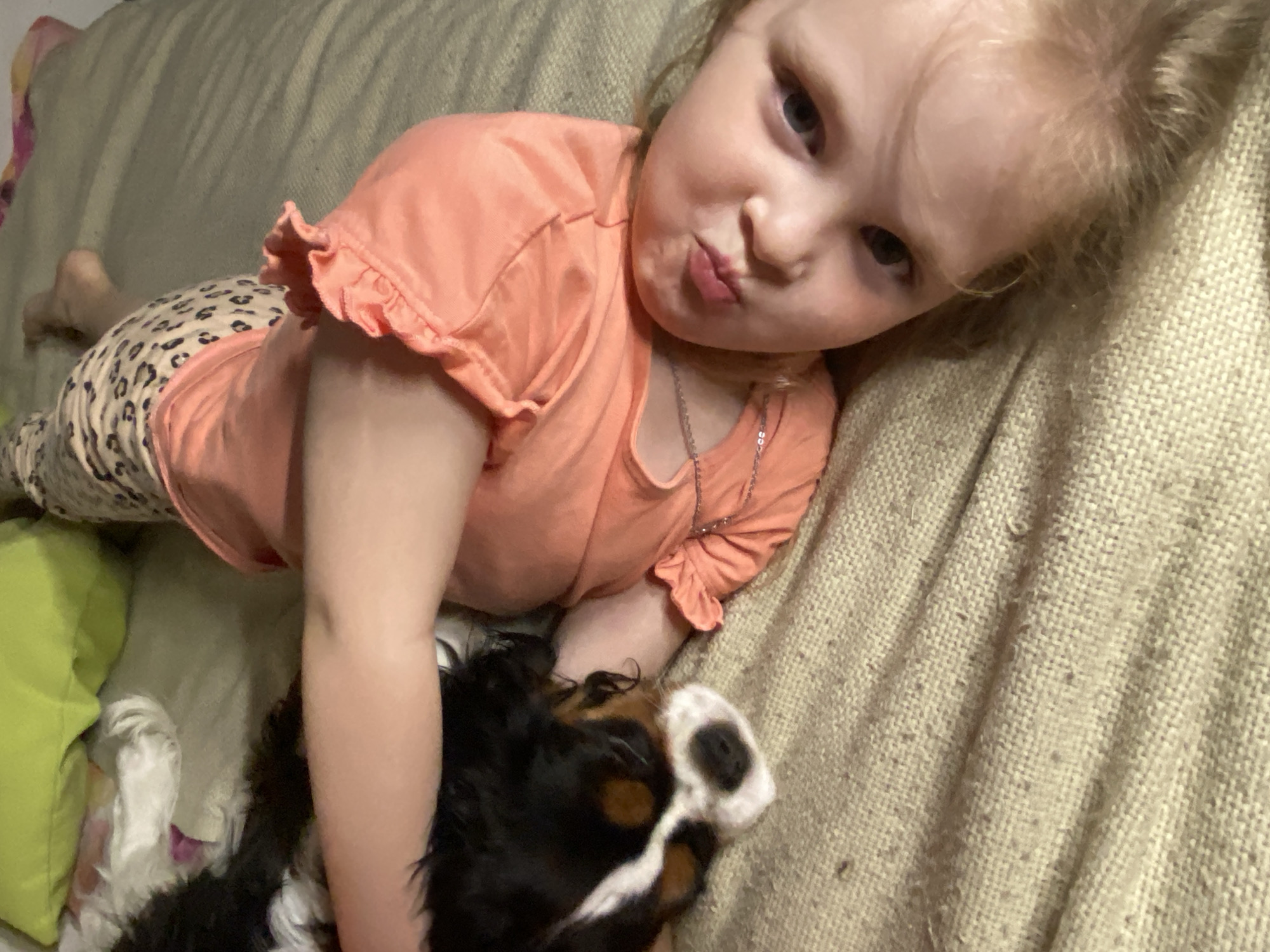 A young child with blonde hair and blue eyes is lying on a light beige couch next to a Cavalier King Charles Spaniel dog. The child is wearing an orange short-sleeved shirt with ruffled edges and patterned leggings. The child is looking at the camera and pursing their lips. The dog is black and brown with a white blaze on its face. The scene is indoors. No information suggests the location is in Votkinsk, Russia.
