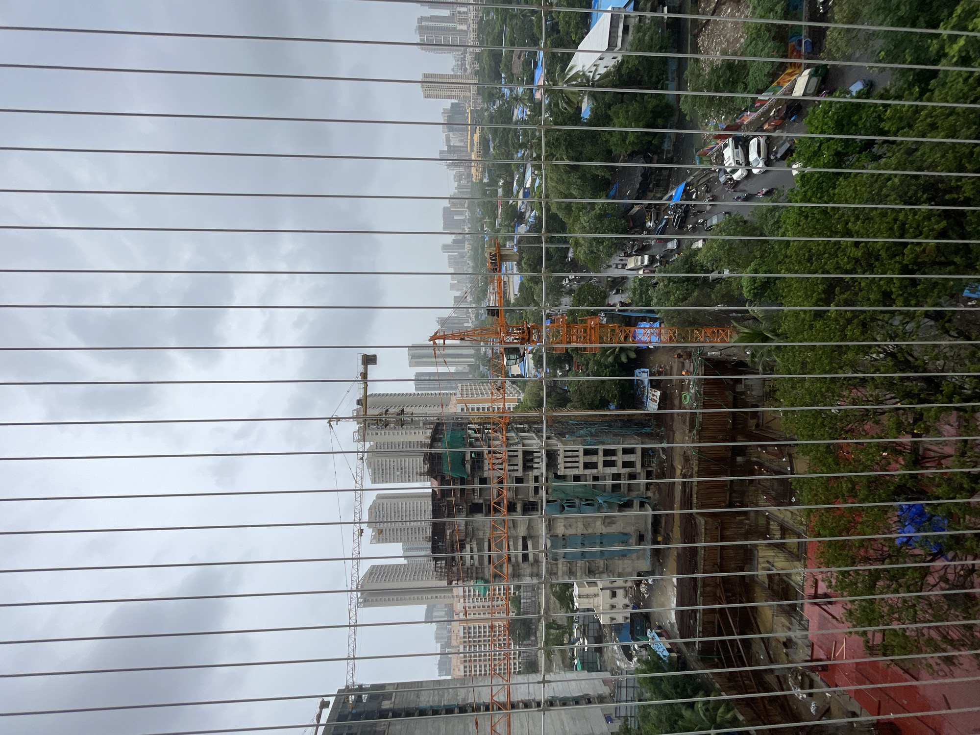 This image, taken from a high vantage point and partially obscured by a wire mesh fence, captures a construction site in Mumbai, India, under a cloudy sky. The scene depicts ongoing construction of a high-rise building, with a prominent orange crane at the center, its arm extended over the partially built structure. Numerous other tall buildings, some completed and others under construction, are visible in the background, suggesting an urban cityscape. Green netting covers parts of the construction, and a large excavated area at the base of one building reveals the foundation work. A street with moderate traffic, including cars and motorcycles, runs through the scene, bordered by lush green trees and smaller buildings. The presence of construction equipment, unfinished structures, and the dense urban environment are indicative of a developing area within Mumbai.