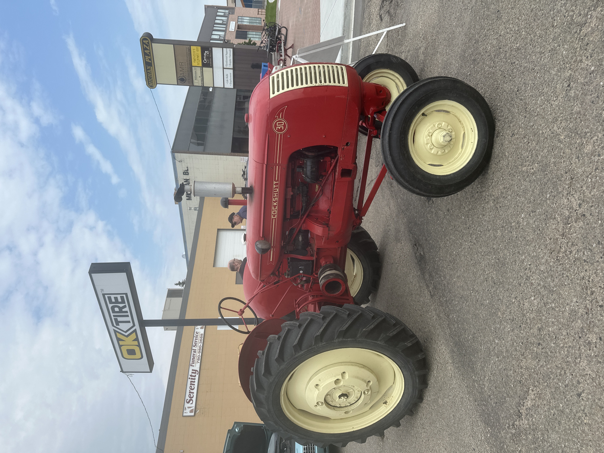 This media file is a vertically oriented, medium-angle shot of a bright red vintage Cockshutt 30 tractor parked on an asphalt surface. The tractor, a prominent object, has yellow rims on its large, black tires and a white and gold grille. Visible text on the tractor includes "COCKSHUTT" and a circular emblem with "30" inside.
The scene is set outdoors, likely during daytime with a cloudy sky. In the background, several buildings are visible. One building has a sign that reads "MICHAN PLAZA" with various business names listed below it, including "AMR," "SVENSEN'S HOLDINGS LTD," and "Century21." Another sign, clearly visible in the foreground, displays "OK TIRE" in large, bold yellow letters. Adjacent to it is a sign for "Serenity Funeral Service" with a phone number.
Several people are present, though their faces are not clearly discernible. One person appears to be looking at the tractor, and another is partially visible near the steering wheel. Bicycles are parked in the background near the buildings.
The overall impression is that of a community event or display, possibly a tractor show or a local gathering, given the presence of vintage machinery and business signage. The weather appears to be overcast. The location is identified by the "OK TIRE" and "MICHAN PLAZA" signs, and the context mentions the City of Leduc, Canada.