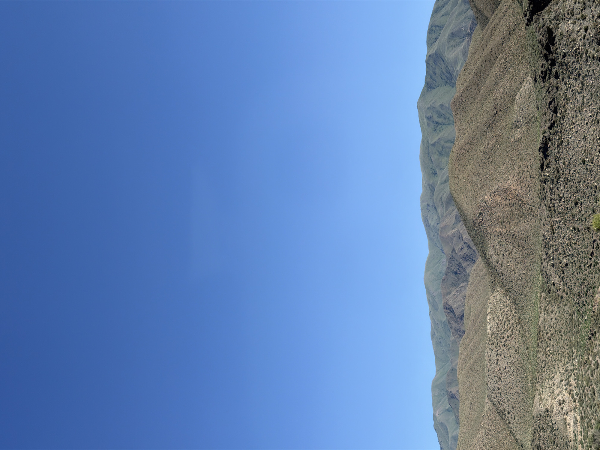 The image depicts a vast, arid mountainous landscape under a clear, bright blue sky. The foreground is dominated by rolling hills covered in sparse, dry vegetation and rocks, with textures ranging from coarse gravel to larger stones. Some areas show faint green patches, suggesting scattered plant life. As the eye moves towards the background, the terrain becomes more rugged, with distinct mountain ranges layered upon one another. These further mountains are a deeper green, their slopes carved with numerous ravines and gullies, indicating erosion. The furthest range appears softer and more distant, almost blending into the horizon. There are no visible people, structures, or immediate signs of human activity. The environment is natural, characterized by its rugged terrain, limited vegetation, and clear atmospheric conditions. The scene is consistent with a high-altitude, dry climate. The location in Kok Moynok Eki, Kyrgyzstan, suggests a remote, natural area.