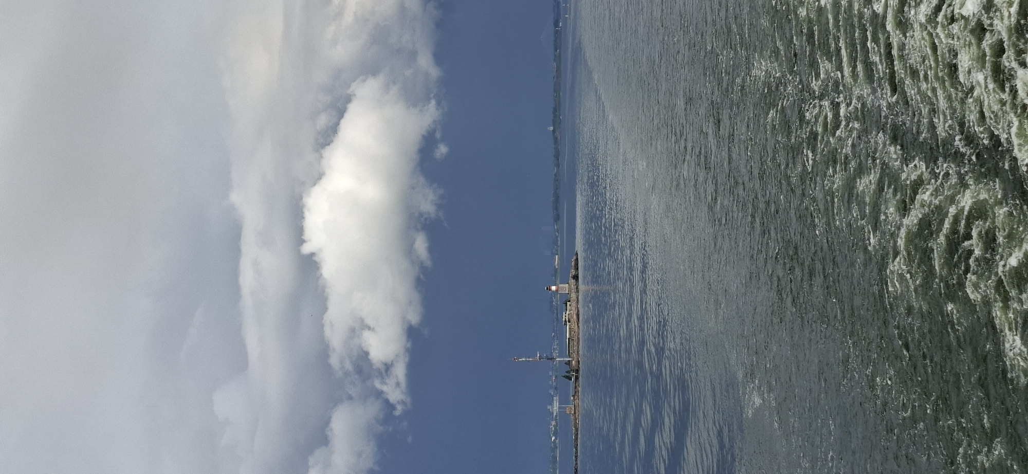 A vertical, medium shot captures a view of the sea with a small island in the distance, featuring a lighthouse and a radio tower. The island is situated to the left of center, with a lighthouse at its southern end, and a tall, slender radio tower situated behind it. The sea, rendered in shades of gray and green, occupies the foreground and middle ground, with white wakes trailing behind the viewer's perspective, indicating a moving vessel. The sky above is a dramatic display of dark gray storm clouds, with a patch of bright white clouds in the center, contrasting with the dark blue expanse of the distant sky. The scene is characteristic of the coastal areas of Helsinki, Finland, possibly near the Suomenlinna sea fortress or other islands in the Helsinki archipelago. There are no people visible in the frame.