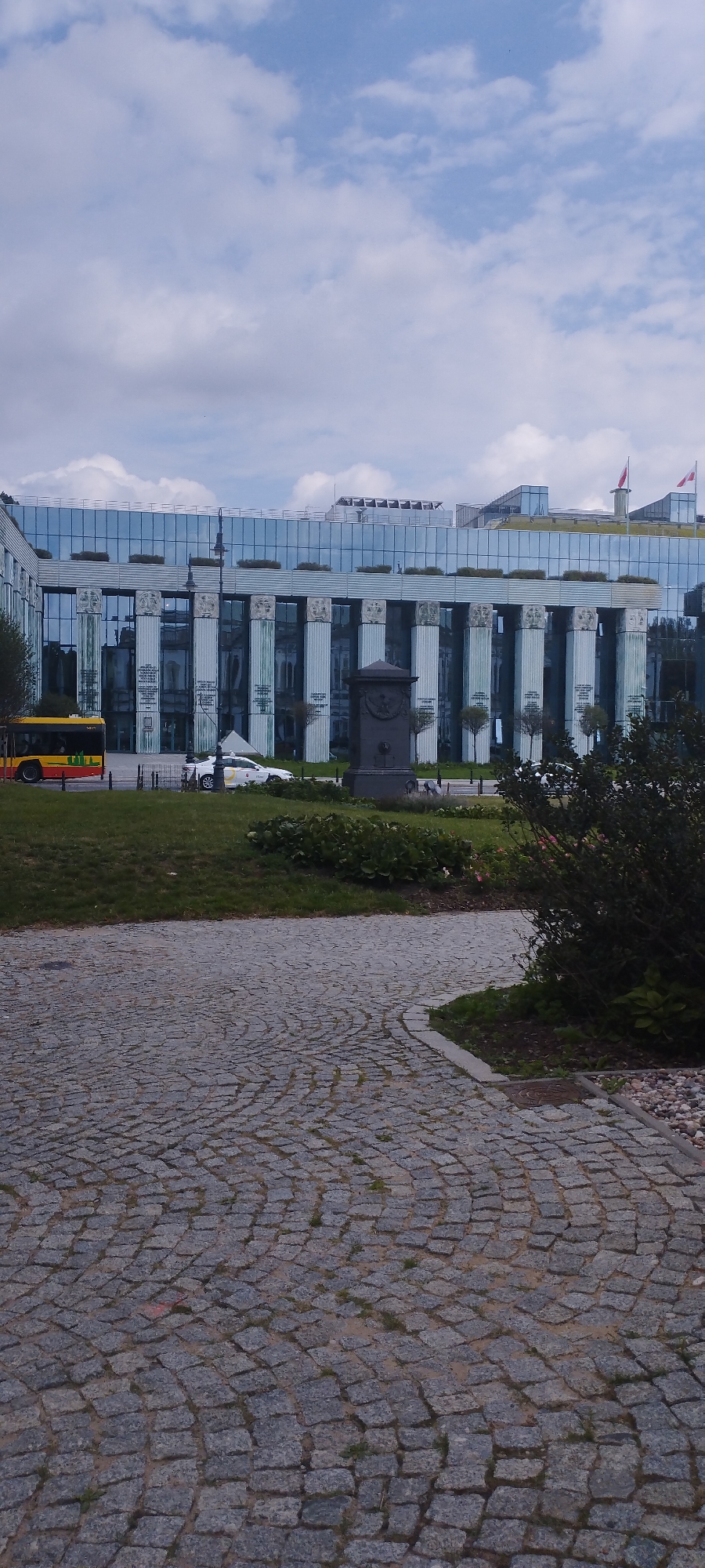 The image shows a modern building with a glass facade and classical columns, possibly the National Bank of Poland in Warsaw, Poland. A yellow and red bus is parked on the left side of the image, and a white car is visible behind it. The foreground is paved with cobblestones. The sky is cloudy.