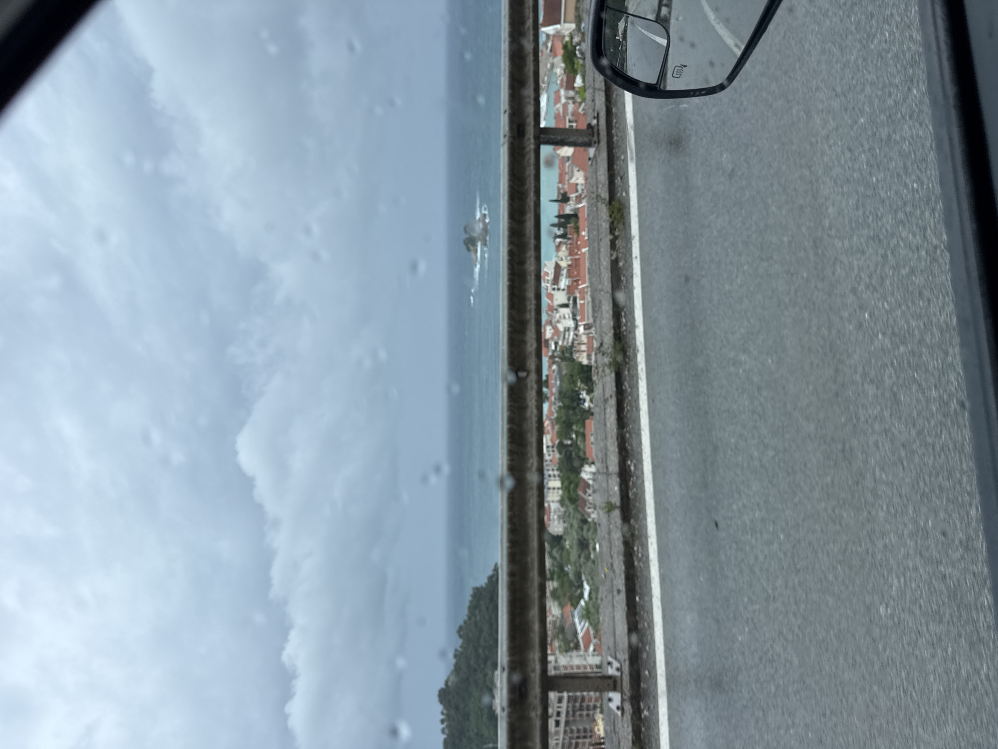 The image shows a view from a vehicle traveling along a road adjacent to the Adriatic Sea in Medinski Krš, Montenegro. The sea, with small rocky islands, is visible on the left, and the town's buildings, distinguished by red-tiled roofs and dense greenery, are situated on the coast. A grey metal guardrail runs parallel to the road, separating it from the seaside view. The road surface is asphalt, marked with a white lane divider. The vehicle's side mirror is visible in the upper right corner of the image, and the front windshield partially obscures the left side of the frame, exhibiting raindrops. The sky is overcast.