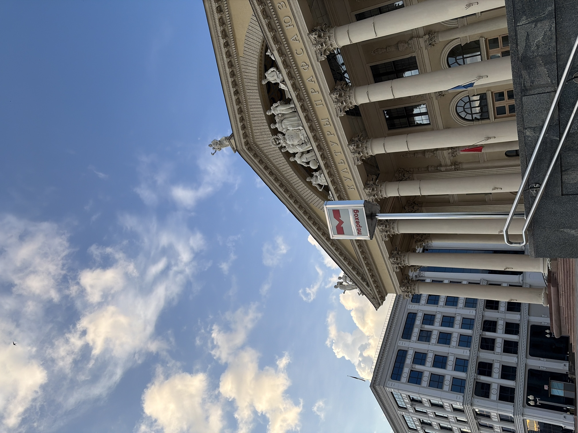 The image shows a wide-angle view of the Palace of Culture of Trade Unions in Minsk, Belarus, on a cloudy day. The palace, a prominent Stalinist Empire style building, is characterized by its grand facade, featuring numerous columns, statues, and ornate carvings. The inscription "КУЛЬТУРЫ ПРАФСАЮЗ" (KUL'TURY PRAFSAYUZ) is visible above the columns, indicating it is the Palace of Culture of Trade Unions.
To the left of the palace, a modern multi-story building with a grid of windows stands. In front of the palace, a sign with the letter "M" in a red circle and the Russian word "переход" (perekhod), meaning "underpass" or "transition," is prominently displayed, suggesting an entrance to an underground passage or metro station.
The sky above is a mix of blue and white clouds, with a small bird visible in the upper left corner. The lighting suggests it is either morning or late afternoon. The foreground shows a stone structure with metal railings, indicating a public area or entrance. There are no people clearly visible in the scene.
