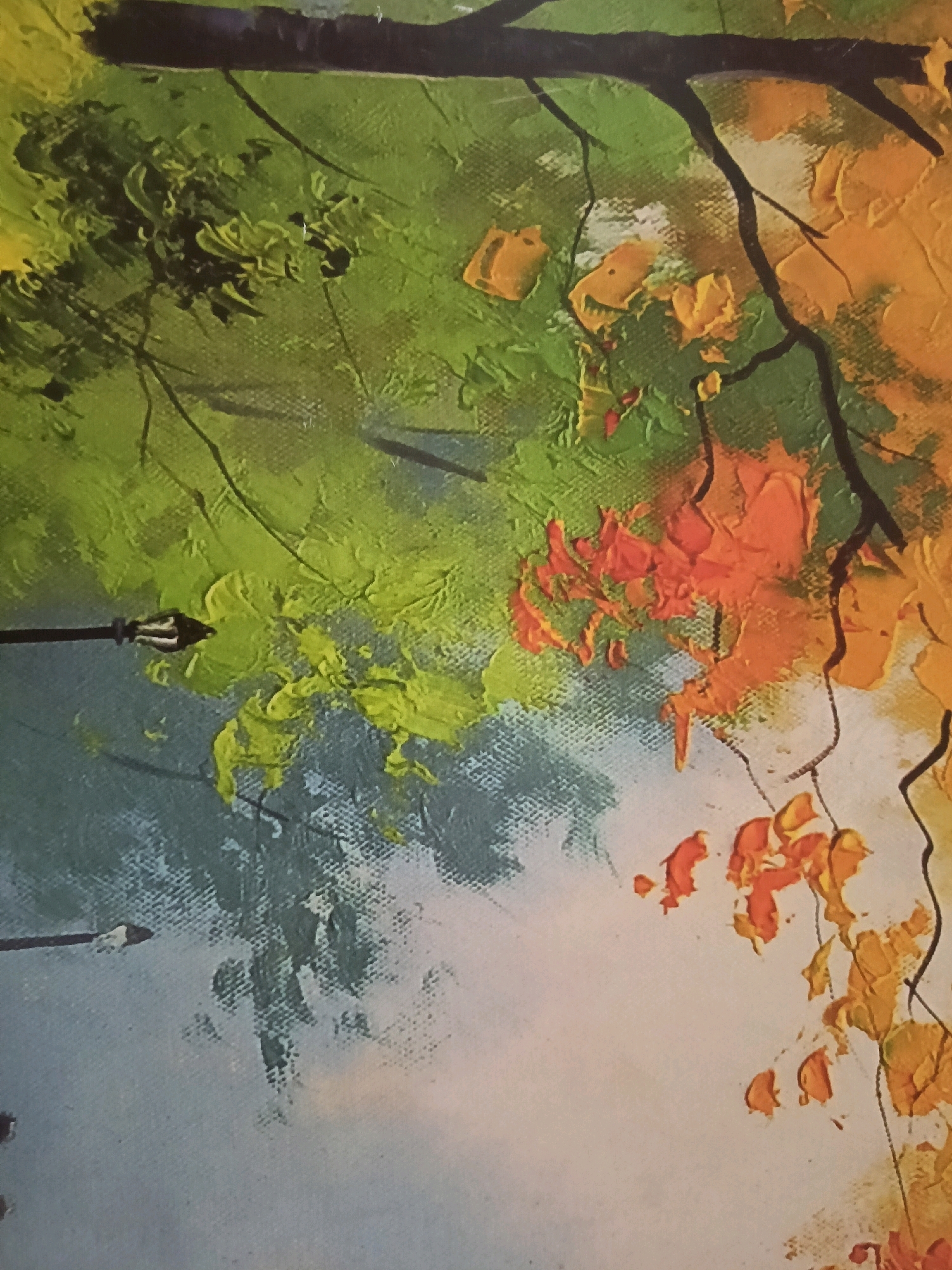 The image is a close-up, cropped, vertical, slightly angled view of an impressionistic painting. The painting depicts a section of a tree with thick, impasto brushstrokes. The upper portion of the image shows a dark, thick branch. Below this, there is a vibrant mix of colors representing foliage, primarily in shades of green, with patches of bright yellow and vibrant orange leaves. The texture of the paint is very prominent, with visible peaks and valleys from the brushstrokes, giving the leaves a textured, almost sculpted appearance. A dark, thin branch extends down from the upper left, and a lamppost, rendered in dark tones with a pointed top, is visible on the left side, emerging from a muted blue-grey background that appears to represent the sky or a distant landscape. The lower portion of the image transitions to a lighter, softer background, possibly indicating a hazy sky or a body of water, with more scattered autumn leaves in yellow and orange. The overall impression is of a scene in nature, likely a park or garden, during autumn, with a focus on the play of light and color through the foliage. There is no visible indication of the city of Karnal, India, or any specific urban features, other than the lamppost, which is a common urban element. The image contains no people.