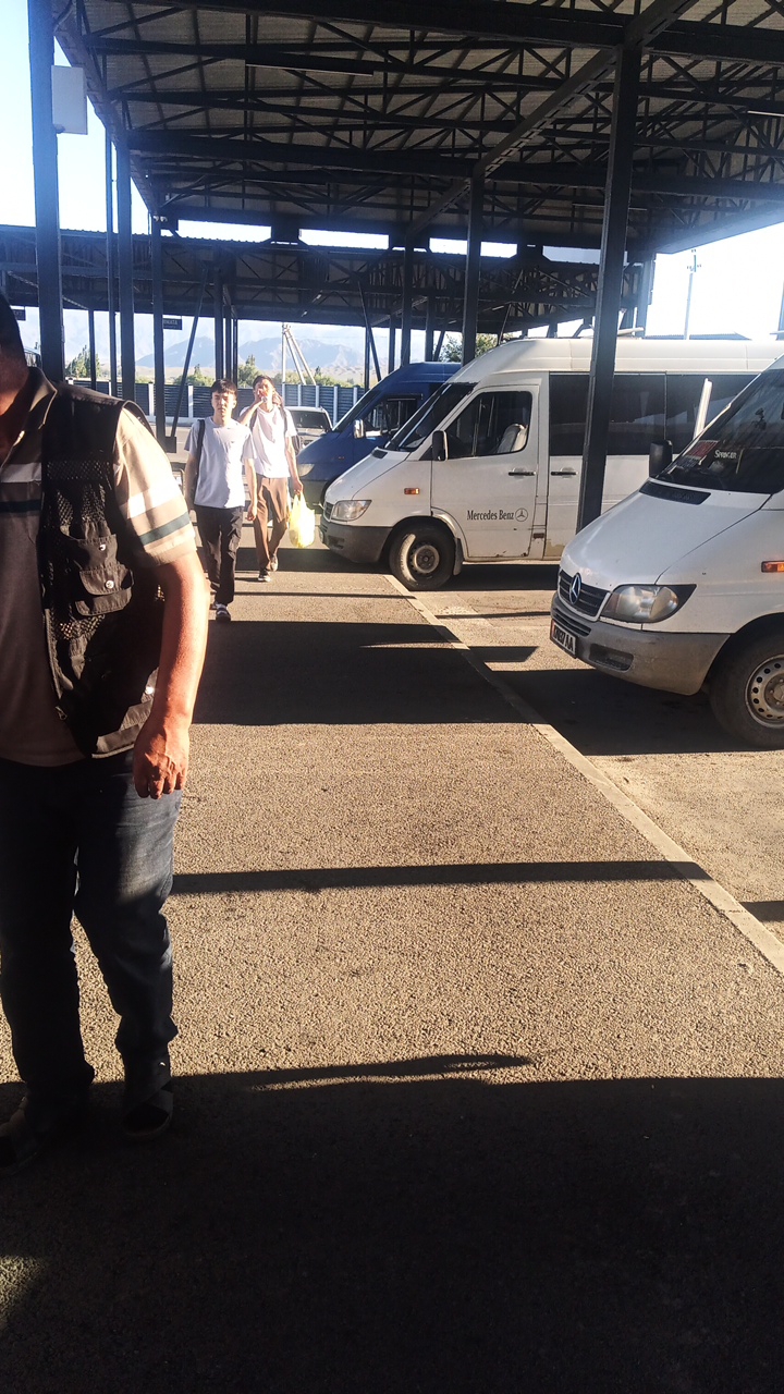 The image depicts a scene at a bus station in Balykchy, Kyrgyzstan. In the foreground, a man wearing a vest over a striped shirt and jeans is partially visible, walking away from the camera. Behind him, two young men, one in a white t-shirt and the other in a white t-shirt and shorts, walk past a line of parked minivans. The minivans, which appear to be white Mercedes-Benz models, are parked under a covered structure with a metal roof and supports. The ground is paved with asphalt. In the background, mountains are visible under a clear sky. The lighting suggests it is daytime, with strong shadows cast by the covered structure.