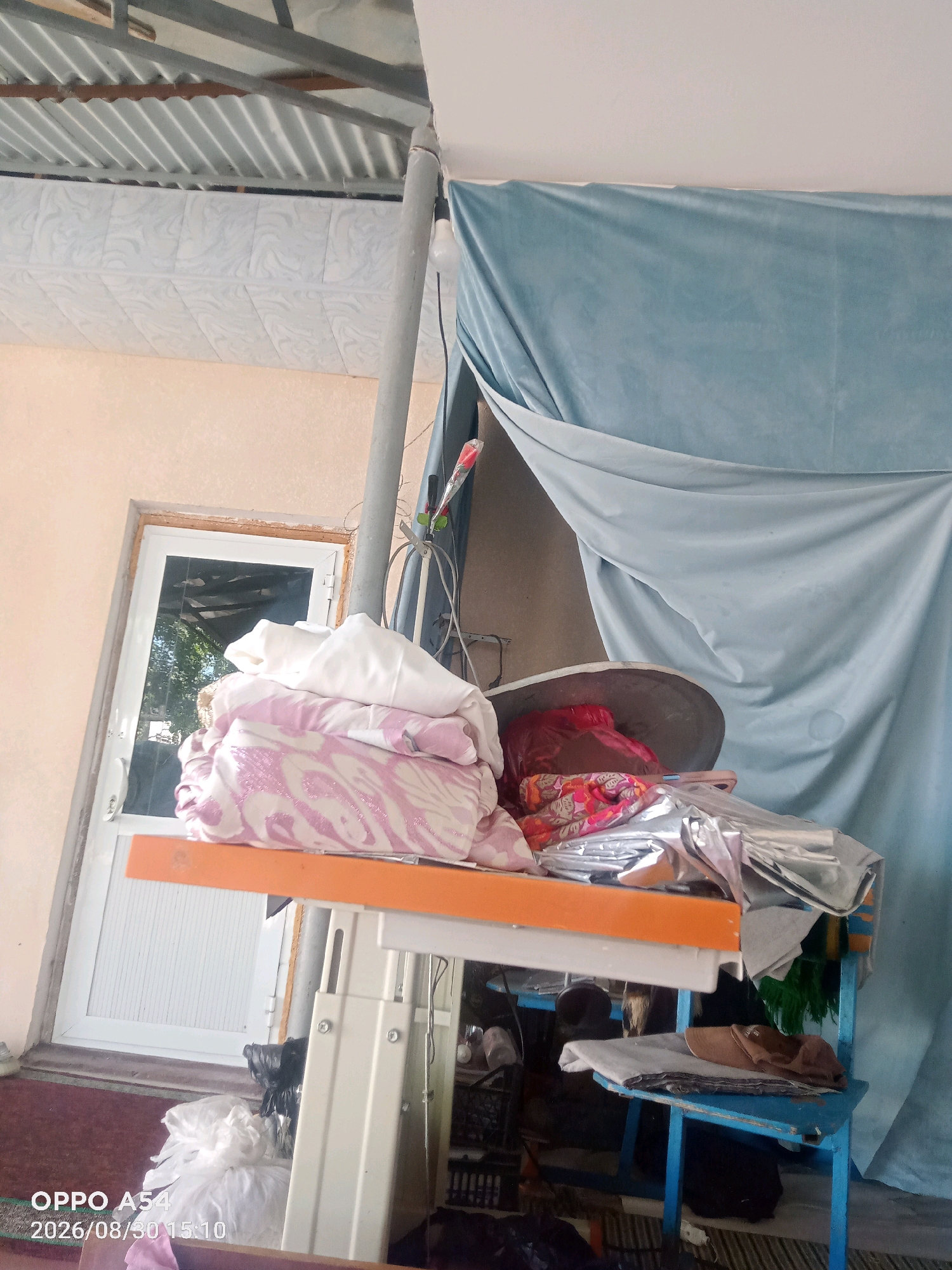 In Zangiata, Uzbekistan, an outdoor scene shows a collection of items on a table and a chair. A stack of white and pink patterned bedding is on an orange shelf. Beside it, a red plastic bag and a patterned fabric are visible. The table itself is a sewing machine base, painted white. Beneath the table, a blue chair holds folded fabric, and a black plastic bag sits on the floor. A white door with a glass pane is to the left, reflecting trees. A large, light blue fabric hangs in the background. The roof above is corrugated metal, supported by beams. A grey pipe runs vertically near the center of the image, and a light bulb hangs from a wire. The timestamp on the image reads "OPPO A54 2026/08/30 15:10".