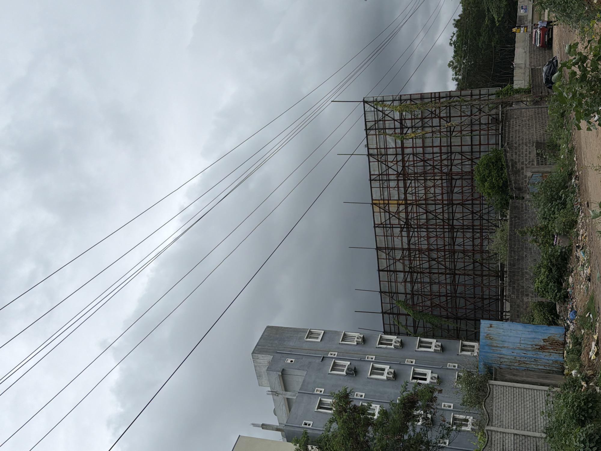 The image captures an overcast day in Shamshabad, India, with a focus on urban infrastructure. In the foreground, a dirt path is littered with various types of debris, including plastic bags and wrappers, indicating a disregard for waste disposal. To the left, a low, grey concrete wall with an ornate pattern runs along the path. Adjacent to it is a tall, rusted, blue metal shed. Behind these elements, a multi-story building stands, its grey facade punctuated by rows of white-framed windows and air conditioning units. To the right of the building, a large, skeletal billboard structure, made of interconnected metal beams, dominates the mid-ground. Green vines are creeping up the structure. Further to the right, a red car is parked on a dirt surface, with two individuals standing nearby. A wider concrete barrier encloses the area in the distance, with lush green trees forming a backdrop. Overhead, a network of dark power lines crisscrosses the grey, cloudy sky. The overall scene suggests an area undergoing development or experiencing neglect, with a juxtaposition of modern construction and the harsh reality of urban waste.