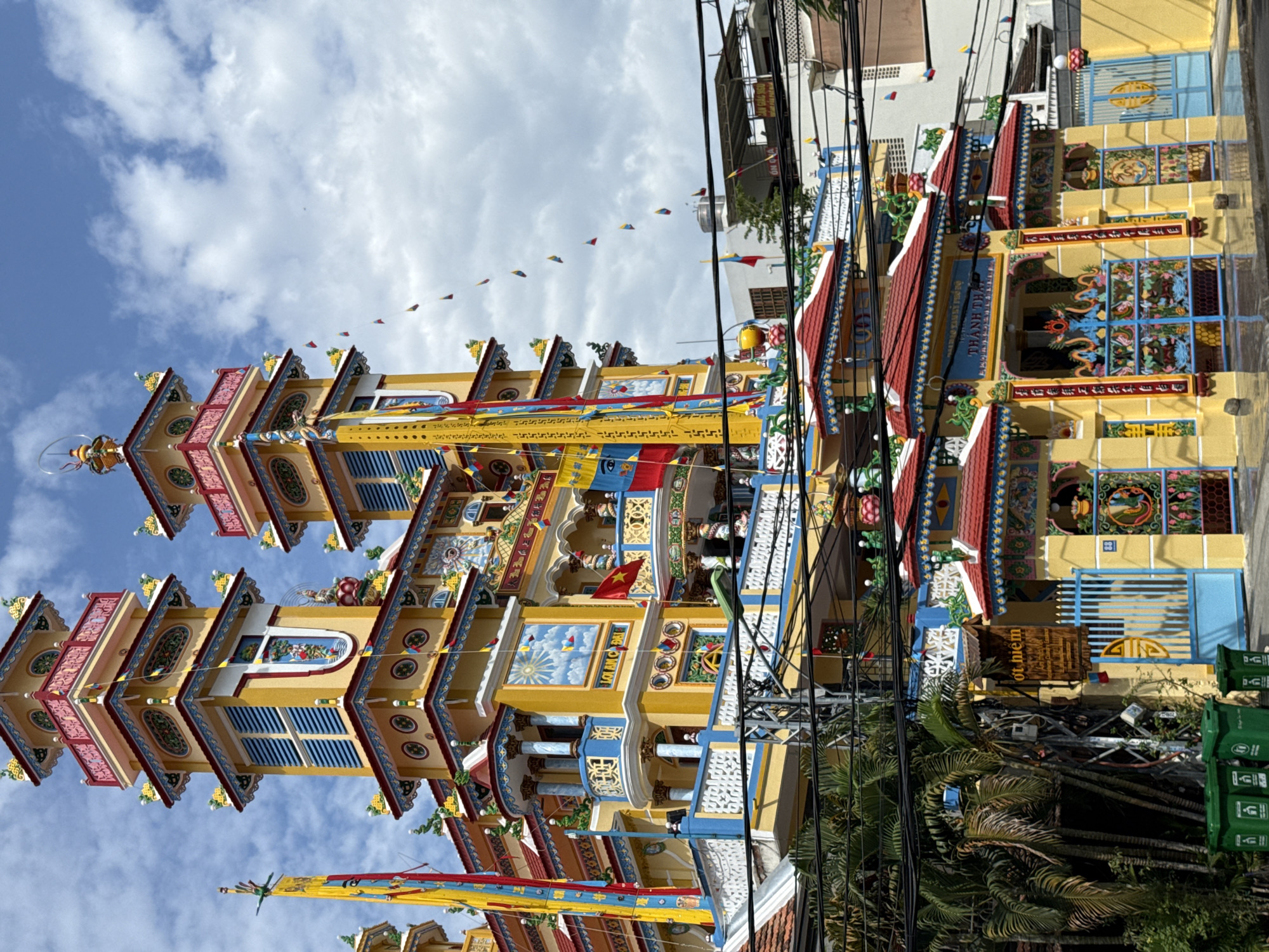 The image shows the exterior of a colorful Buddhist temple in Nha Trang, Vietnam. The temple features multiple ornate towers with yellow and red accents, adorned with intricate carvings and decorative elements. Several flags, including Vietnamese flags and smaller triangular pennants, are displayed prominently. The foreground is dominated by a street with visible power lines and palm trees, suggesting an urban environment. The temple has a vibrant yellow facade with blue and red detailing, and elaborate gates with dragon motifs. The overall scene depicts a bright, sunny day with some clouds in the sky, highlighting the vivid colors and architectural style of the temple.