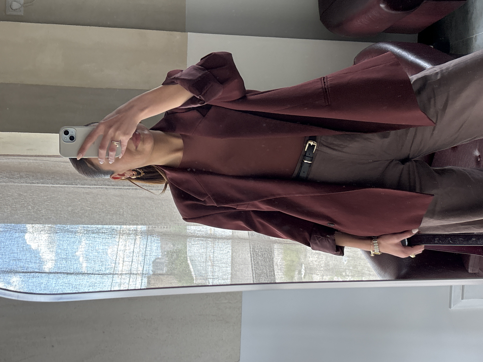 A woman is standing indoors, reflected in a mirror. She is holding up a white iPhone in her right hand, presumably taking a selfie. She is wearing a maroon outfit, which includes a top, pants belted at the waist with a black belt with gold buckle, and a blazer or jacket. She is also wearing a gold watch, a ring, and earrings. The background visible through the window shows a view of trees and the sky. The room appears to be neutral-colored, with an armchair or chair partly visible in the upper right. The location is unknown and there is no way to determine whether the location is in Yerevan, Armenia.