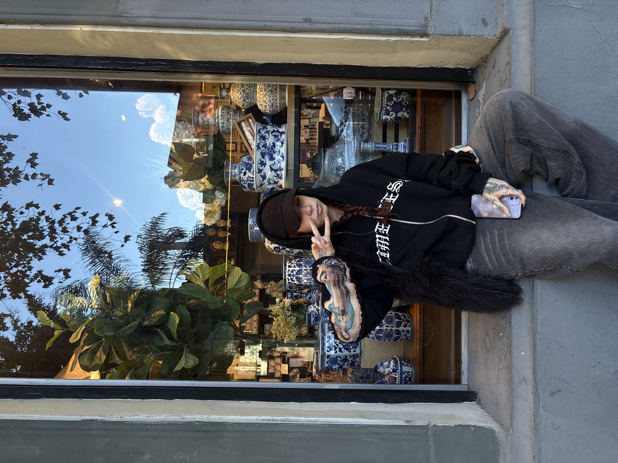 A young person, likely a woman, is seated outdoors, leaning against a grey concrete wall or ledge next to a large storefront window. She is wearing a black zippered hoodie with stylized white text on the chest/sleeve, a dark baseball cap, and baggy dark grey pants. The hoodie features a black, fur-like trim around the neck. She has long, dark braided hair, a visible nose piercing, and tattoos on her left forearm (depicting a figure, possibly religious) and on her right hand. Her left hand is raised, forming a peace sign, while her right hand rests on her lap, holding a light purple smartphone with long, light-colored fingernails.
The storefront window behind her displays a collection of blue and white ceramic vases and decorative items, suggesting an antique or home decor shop. White artificial floral arrangements are also visible within the display. The window strongly reflects the bright blue sky with white clouds, along with the silhouettes of tree foliage, including broad leaves and palm fronds, indicating a sunny daytime setting. A large green potted plant is also visible inside the store, reflecting in the lower left portion of the window. The overall scene depicts an urban setting with a building facade. The person appears relaxed and playful, directly facing the camera. The context given for the image location is Buenos Aires, Argentina.