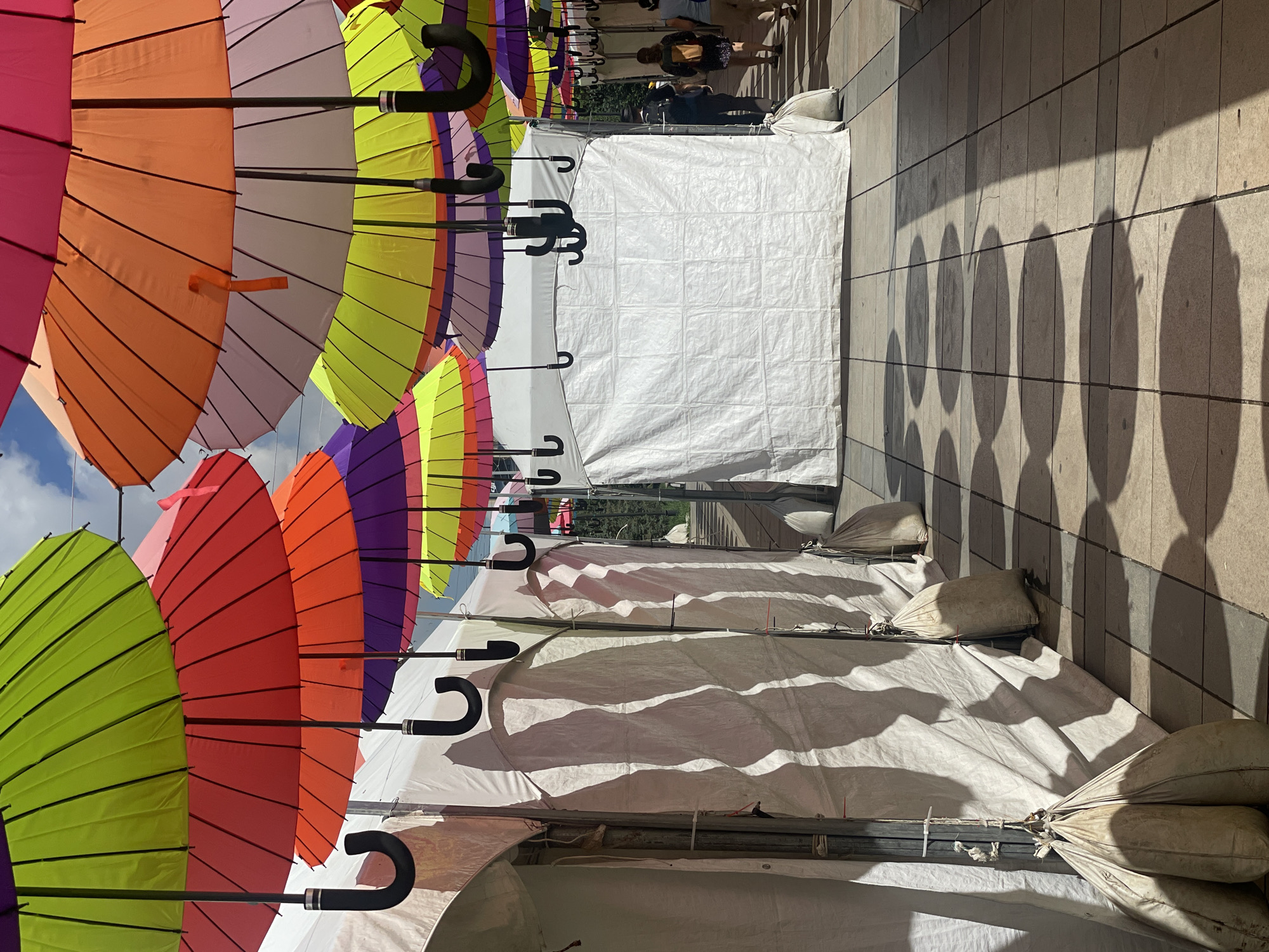 The image depicts a vibrant outdoor market or festival scene, with numerous colorful umbrellas suspended overhead. The umbrellas are a mix of bright hues, including lime green, red, purple, orange, and pink, creating a festive canopy.
In the foreground, white tents line the walkway, casting long, dramatic shadows on the tiled ground. These tents appear to be part of market stalls or event booths. Sandbags are visible, used to anchor the tents.
In the middle ground, a larger white tent structure is present. Beyond this, a walkway is visible, with a few individuals walking away from the camera. One person is wearing a dark shirt and backpack, while another has on a lighter-colored top and a tan bag.
The background shows a glimpse of trees and a bright sky, suggesting an outdoor setting on a sunny day. The overall impression is one of a lively public gathering, likely a temporary event or market.
While the image displays a lively outdoor setting with stalls and umbrellas, there is no specific visual information within the image that allows for the determination of its location in Ulaanbaatar, Mongolia.