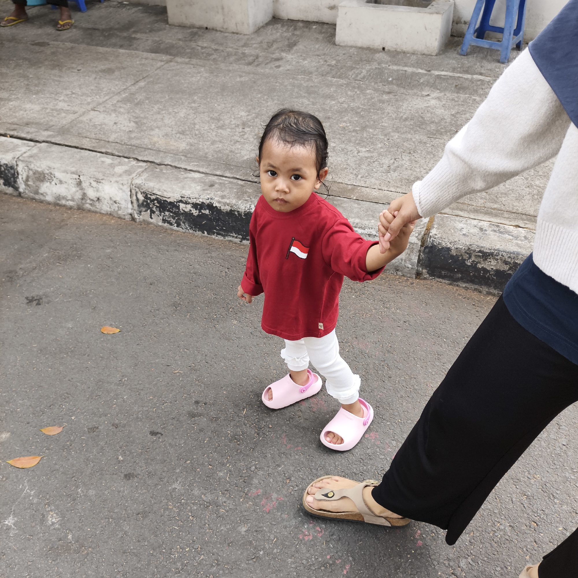 A toddler wearing a red long-sleeved shirt with a red and white flag emblem on the left chest, white leggings with ruffled ankles, and pink slip-on sandals is being led by the hand by an adult wearing a cream-colored long-sleeved top with a dark blue band and black pants. They are walking on an asphalt road next to a concrete sidewalk with a black and white striped curb. The scene appears to be outdoors, possibly in a residential or public area of South Jakarta, Indonesia, as indicated by the presence of the Indonesian flag on the child's shirt. Two fallen leaves are visible on the asphalt in the foreground. In the background, part of a white building and a blue plastic stool can be seen. The child is looking directly at the camera with a serious expression.
