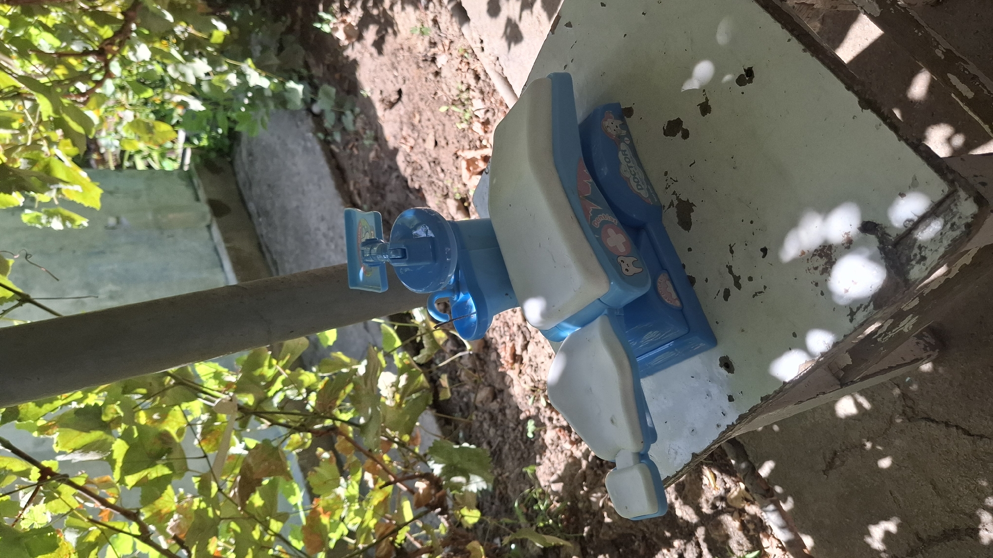 A blue and white toy dental chair sits on a weathered, pale green surface. The toy dental chair features a seat, armrests, a headrest, and a small basin. There are cartoonish tooth and medical cross graphics on the side of the chair, along with the word "DOCTOR". The surface the toy is resting on appears to be the top of an old table or bench, with chipped paint and dark stains. The background shows a natural, outdoor environment with green foliage and soil. A thick, gray pole is visible behind the toy. Sunlight casts shadows across the scene. The location is Шұбарсу, Kazakhstan.