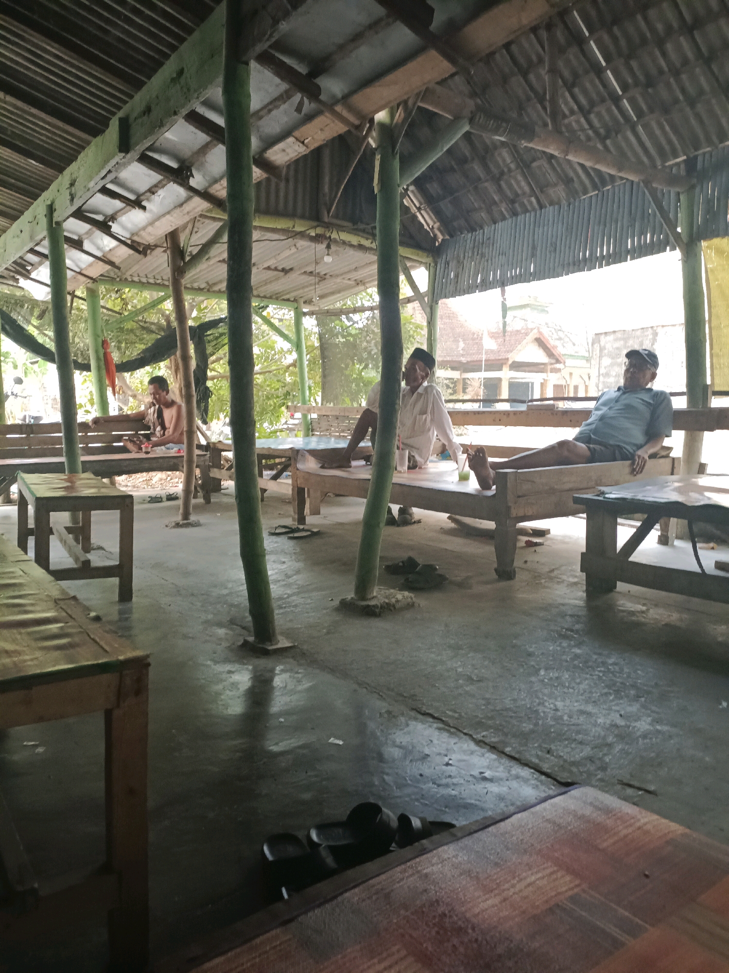 In Rancang Kencono, Indonesia, an outdoor seating area is depicted under a corrugated metal roof supported by wooden and bamboo poles. The floor is a concrete surface, wet in places. Three men are present. In the background, a shirtless man sits on a wooden bench, holding a small object. In the mid-ground, to the left, a man in a white shirt and black cap is seated at a table with a drink. To his right, a man in a blue shirt and a black baseball cap reclines on a bench, with his legs extended. Several wooden tables and benches are arranged throughout the space. In the foreground, parts of wooden tables are visible, with a woven mat on one. Sandals are scattered on the floor near the benches. The environment appears to be a casual, open-air gathering place.