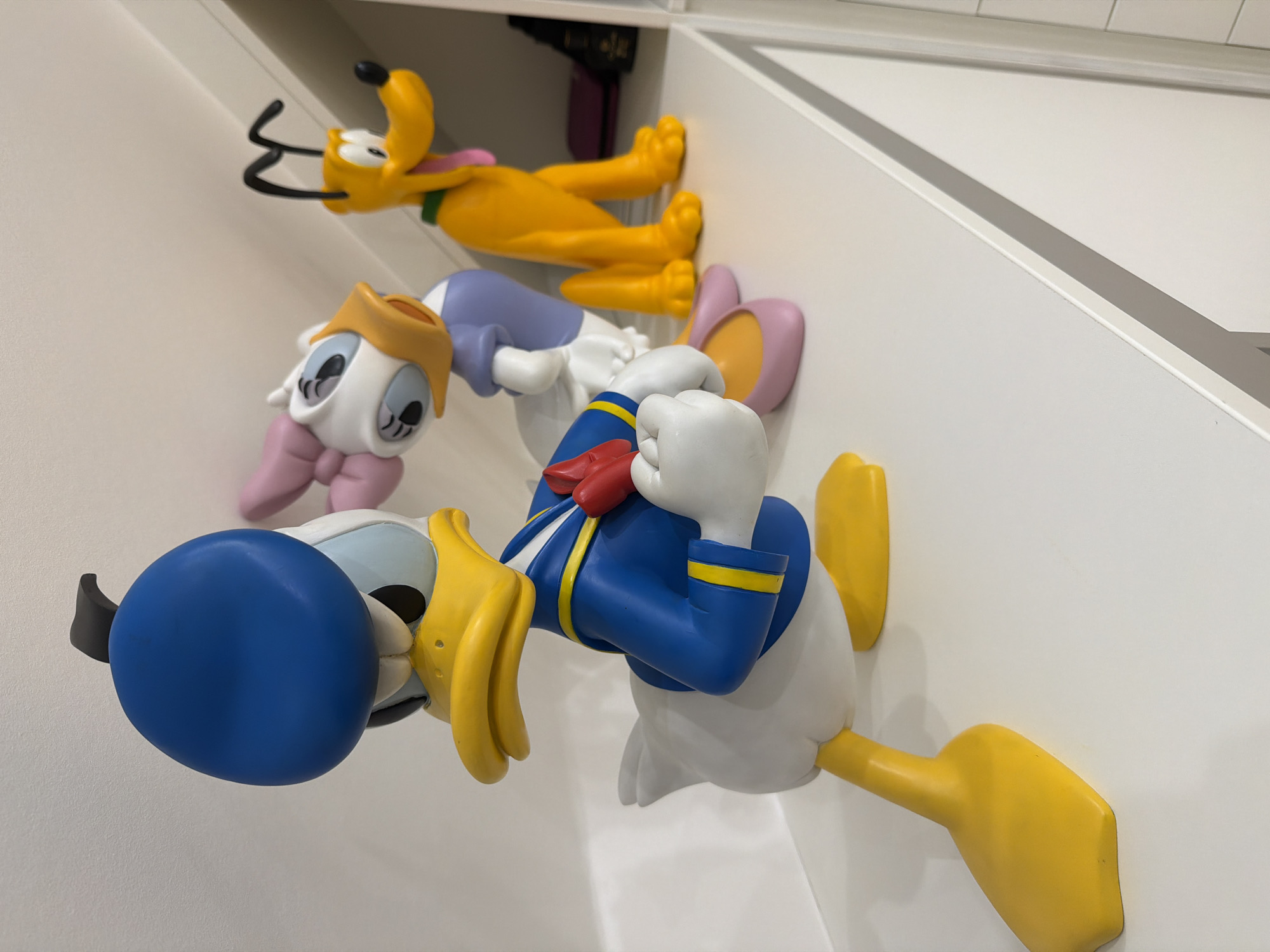 The image shows a set of Disney character sculptures mounted on a white wall, consisting of Donald Duck, Daisy Duck, and Pluto. Donald Duck is in the foreground with a blue sailor hat, blue jacket with gold trim, and a red bow tie. Daisy Duck is behind him, wearing a lilac dress, pink shoes, and a golden visor on her head. Pluto is positioned at the top of the arrangement, lying on his stomach. The sculptures are displayed on an angled wall, with white shelving visible at the top. The photo was taken in Pétange, Luxembourg.