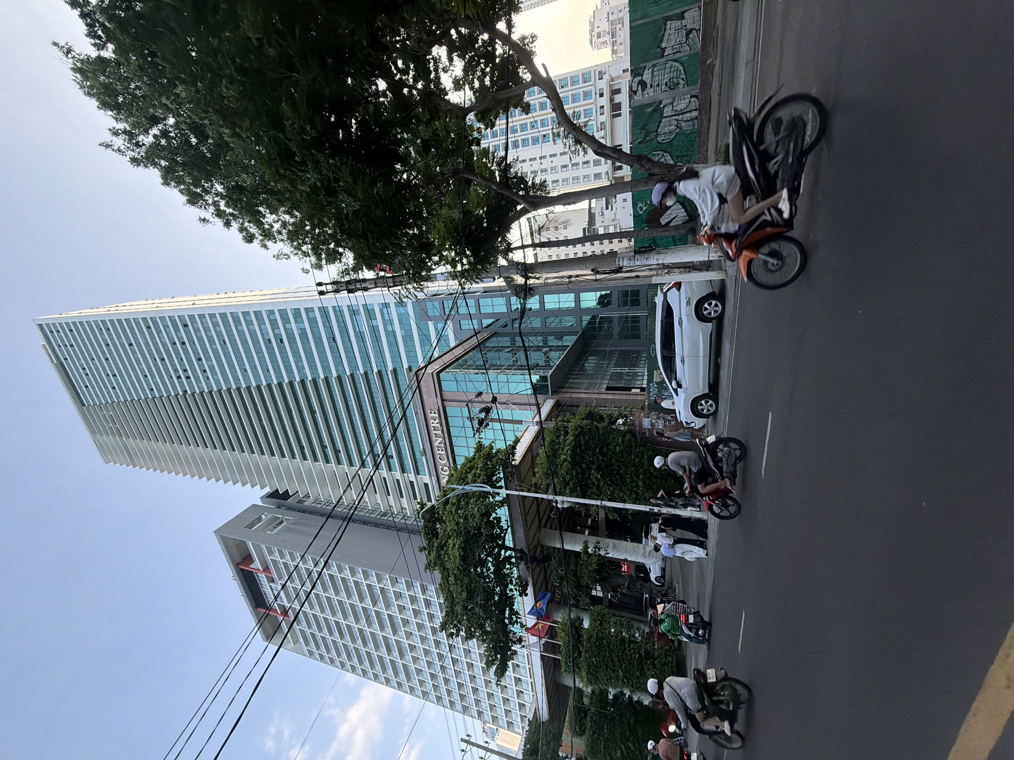 The image depicts an urban street scene in Nha Trang Ward, Vietnam, specifically on Tran Phu Street in front of the Gold Coast Condotel (address: 25 Tran Phu, Loc Tho, Nha Trang, Khanh Hoa, Vietnam).
The central focus is a very tall, slender skyscraper with a facade composed of light blue-green reflective glass panels and prominent white horizontal balcony-like structures. To its left, another tall building exhibits a contrasting gray exterior with a grid pattern of white balconies and red structural elements near its apex. At the street level, between these two towers, is a modern multi-story glass-fronted building segment displaying the signage "G CENTRE."
On the asphalt roadway, several motorcycles are in transit. A rider in a white and black striped shirt is visible on a dark motorcycle on the left, followed by another rider in a green shirt. On the right, a person wearing a white long-sleeved top and a purple cap operates an orange and black scooter. Multiple pedestrians are gathered on the sidewalk near the entrance of the "G CENTRE" building on the left. A white SUV is either parked or moving slowly near the curb on the left. The street features painted white dashed and yellow solid lines.
The environment includes mature green trees lining the street, partially obscuring parts of the buildings and power poles. A dense network of black power lines spans across the upper portion of the frame. On the right side of the road, a green barrier, possibly for construction, is visible and covered with graffiti. A tall utility pole laden with cables stands near the right edge of the central building. The sky overhead is a clear light blue with minimal cloud cover.
