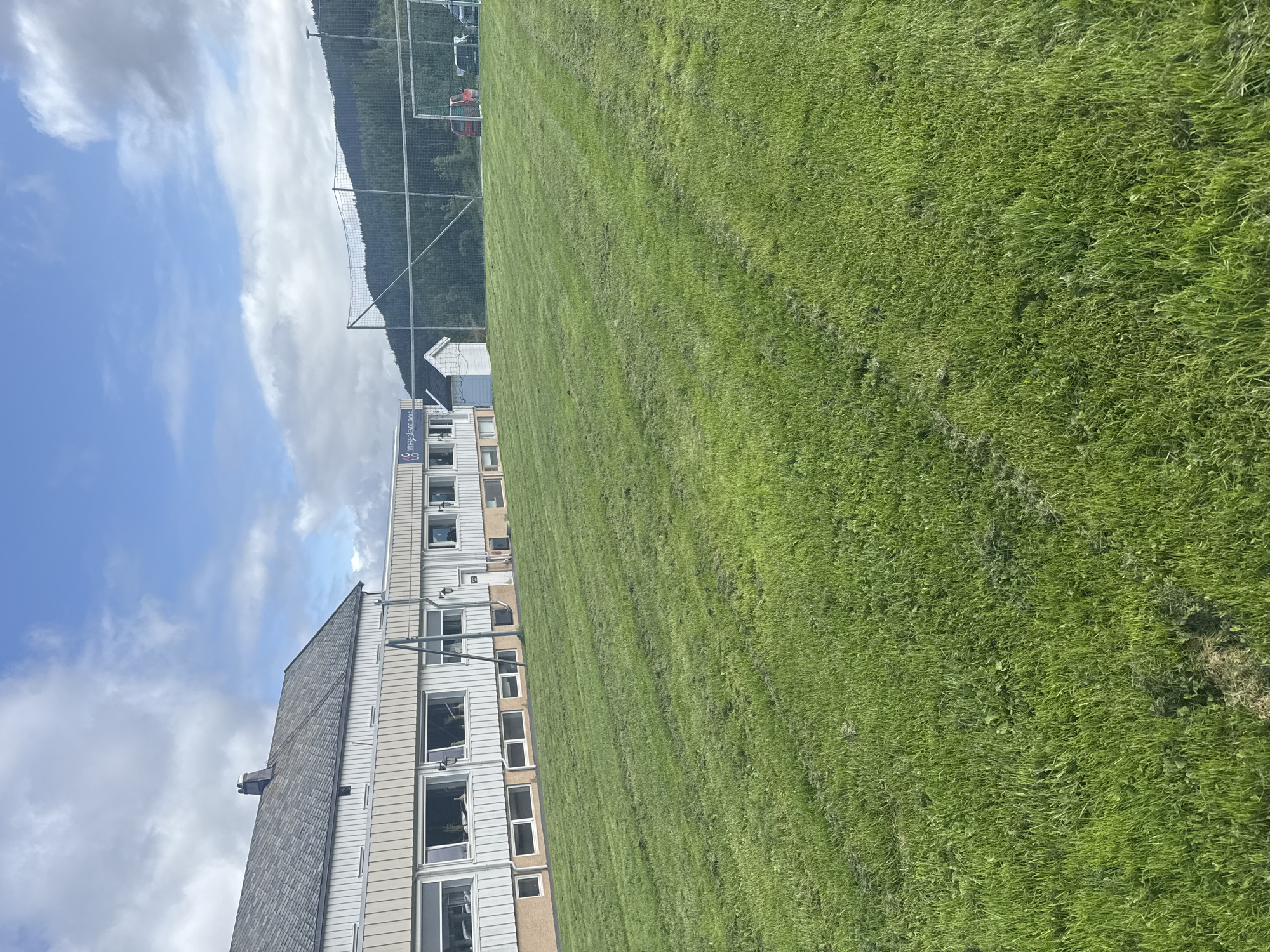 The image depicts a wide, green, freshly cut grass field in front of a building and a sports area. The building, which appears to be a school or community center, has a light-colored exterior with a dark gray roof. A sign with text "AG VIRKEGÅRDEN SKOLE" is visible on the building. To the right of the building, there's a small white shed. Beyond the building and to the right, a sports field with a net and goalposts is visible, along with a few parked vehicles. The background features a lush green forest leading up to a mountain range under a partly cloudy sky. This scene is likely located in Skatval, Norway, as indicated by the sign on the building. There are no people visible in the image.