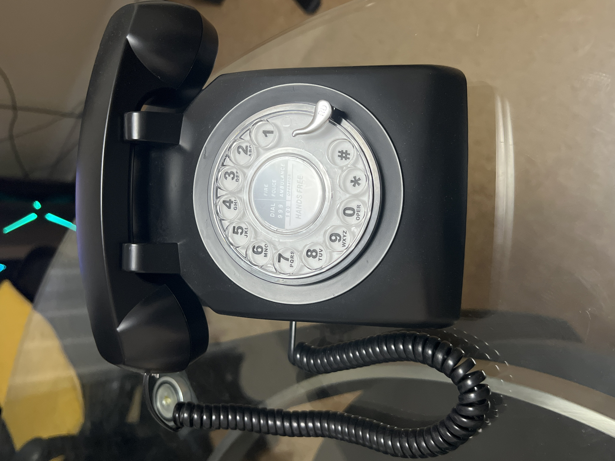 A black, rotary-dial telephone sits on a glass surface. The dial face includes numbers 1-9, 0, #, and *. The text "DIAL FIRE POLICE AMBULANCE 999 100 OPERATOR HANDS FREE" is visible at the center of the dial. The phone's coiled cord extends from the base.
The location cannot be determined from the image.