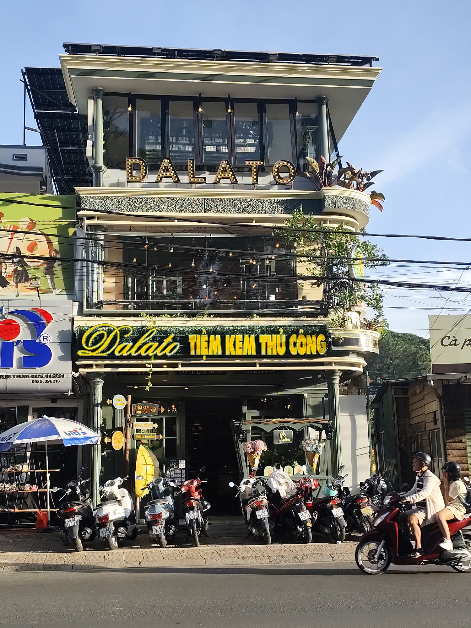 The image depicts the exterior of the Dalato ice cream shop in Da Lat, Vietnam, on a sunny day. The building has a modern design with multiple stories and large windows. A prominent neon sign on the second floor reads "Dalato Tiệm Kem Thủ Công," indicating it's a handmade ice cream shop. Several motorcycles are parked in front of the shop, and two people are riding a motorcycle on the street in the foreground. An umbrella is set up to the left of the entrance, possibly for shade. Electrical wires are strung across the facade of the building. The sky is clear and blue.