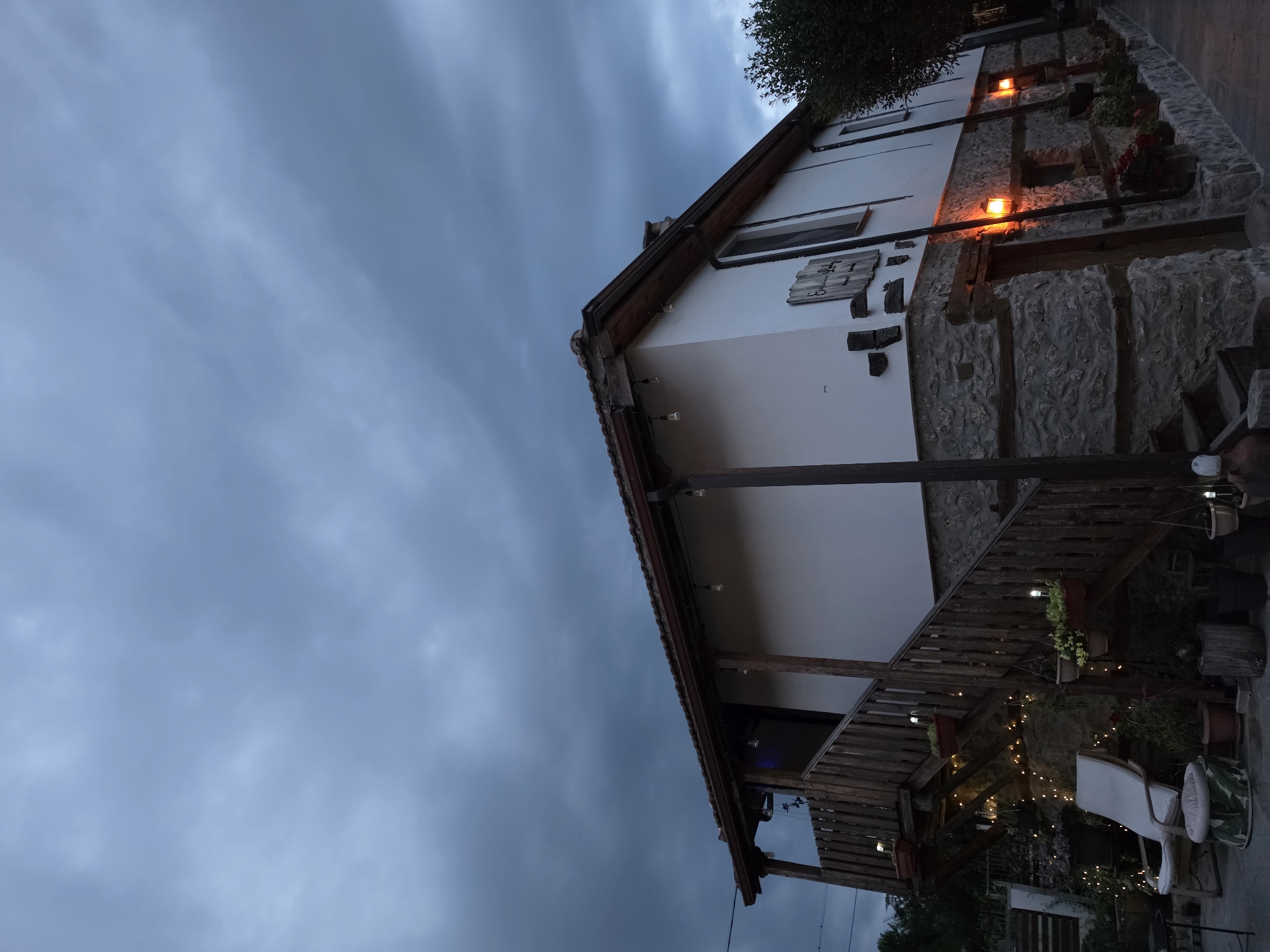 An exterior shot shows a two-story stone and stucco building at dusk. The sky above is filled with dark, heavy clouds. The building features a wooden porch with an outdoor staircase leading to the upper level, adorned with string lights. The lower level has a stone facade and several windows and doors, with orange outdoor lights illuminating the area. There are potted plants and garden decor around the entrance. To the right of the building, a tree is visible. The scene appears to be in a rural or semi-rural setting, possibly a guesthouse or restaurant.