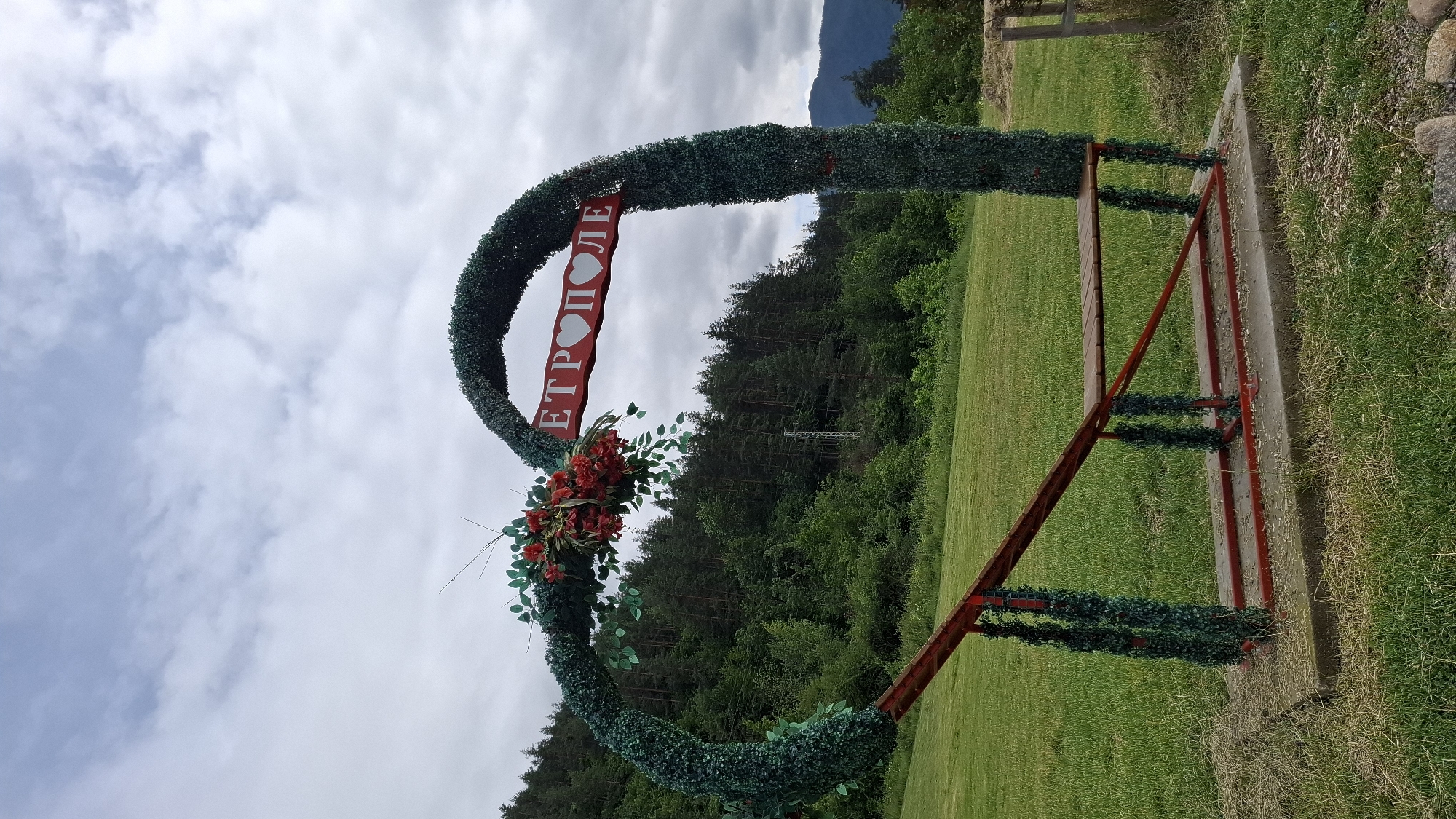 This is a medium shot, eye-level, outdoor photograph of a heart-shaped archway with a bench underneath. The archway is covered in green foliage and decorated with red flowers. A red sign with white lettering and a heart symbol reads "ЕТРОПОЛЕ". The archway is situated in a grassy area with a backdrop of a dense forest. The sky is overcast with scattered clouds. The bench is made of wood and painted red. The entire structure is placed on a concrete base in a rural setting outside of the city of Etropole, Bulgaria.