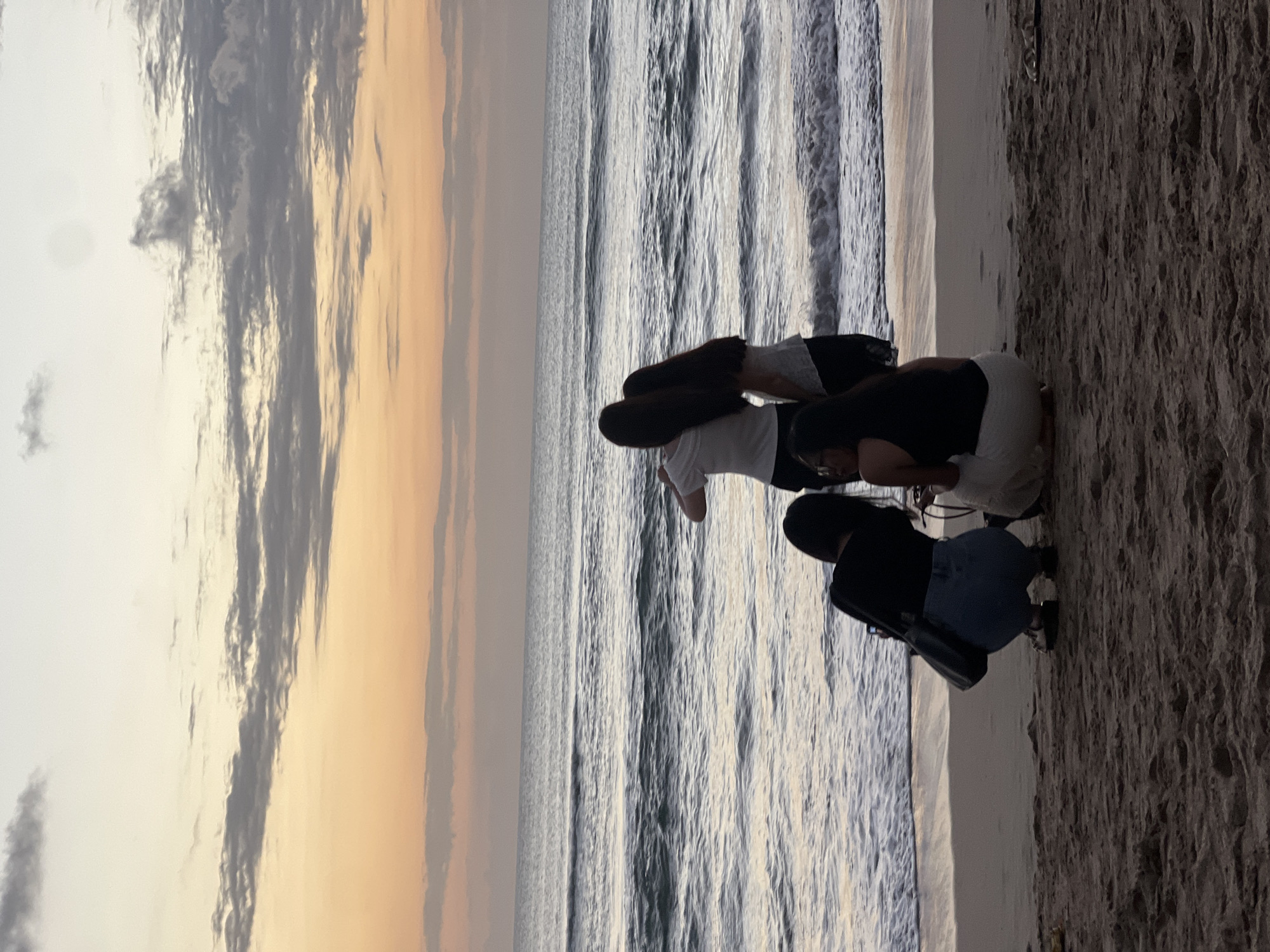 Four people are visible on a sandy beach in Kuta, Indonesia, facing the ocean. The sky is a gradient of pale yellow and orange, with scattered clouds, suggesting either sunrise or sunset. The ocean has moderate waves breaking on the shore. Two people are standing with their backs to the camera, facing the sea, and two people are crouched on the sand. One of the crouched individuals is wearing a black top and blue jeans, and appears to be taking a photo with a phone. The other crouched individual wears a black tank top and white pants. The sand is textured with footprints and debris.