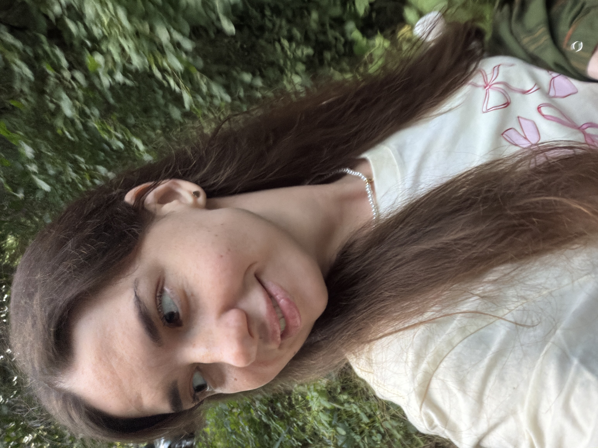 A medium close-up shot captures a young woman with brown hair and light skin, wearing a cream-colored t-shirt with pink bow prints, and a pearl necklace. She is looking to her left, with a slight smile on her lips. The background consists of lush green foliage, suggesting an outdoor setting in a park or garden. The scene appears to be in the village of Kmytiv, Ukraine, as indicated by external information. There are no other people or objects of significance in the frame, and the overall atmosphere is casual and relaxed.
