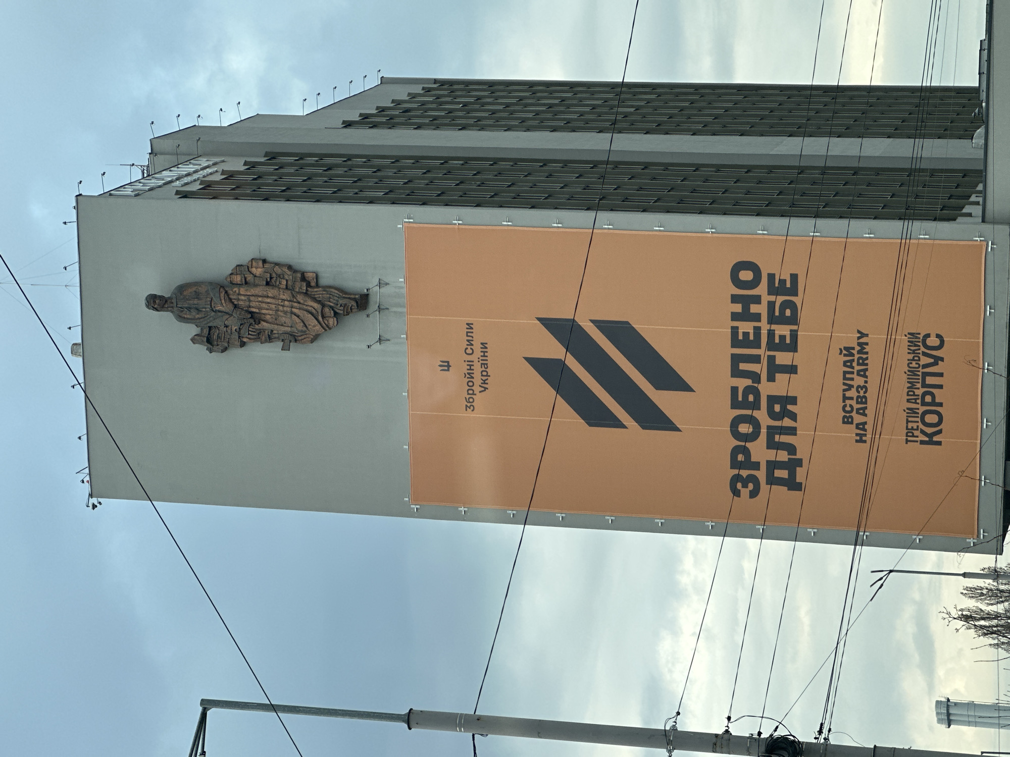 The image shows the building of the Ministry of Defense of Ukraine in Kyiv with a large advertisement for recruitment to the Ukrainian army placed on the facade. The advertisement is in Ukrainian, including phrases like "Made for you" and "Join the AB3.ARMY - Third Army Corps". There is also a bronze sculpture depicting a seated figure on the building. The sky is overcast. The image is taken from a low angle, showcasing power lines in the foreground.