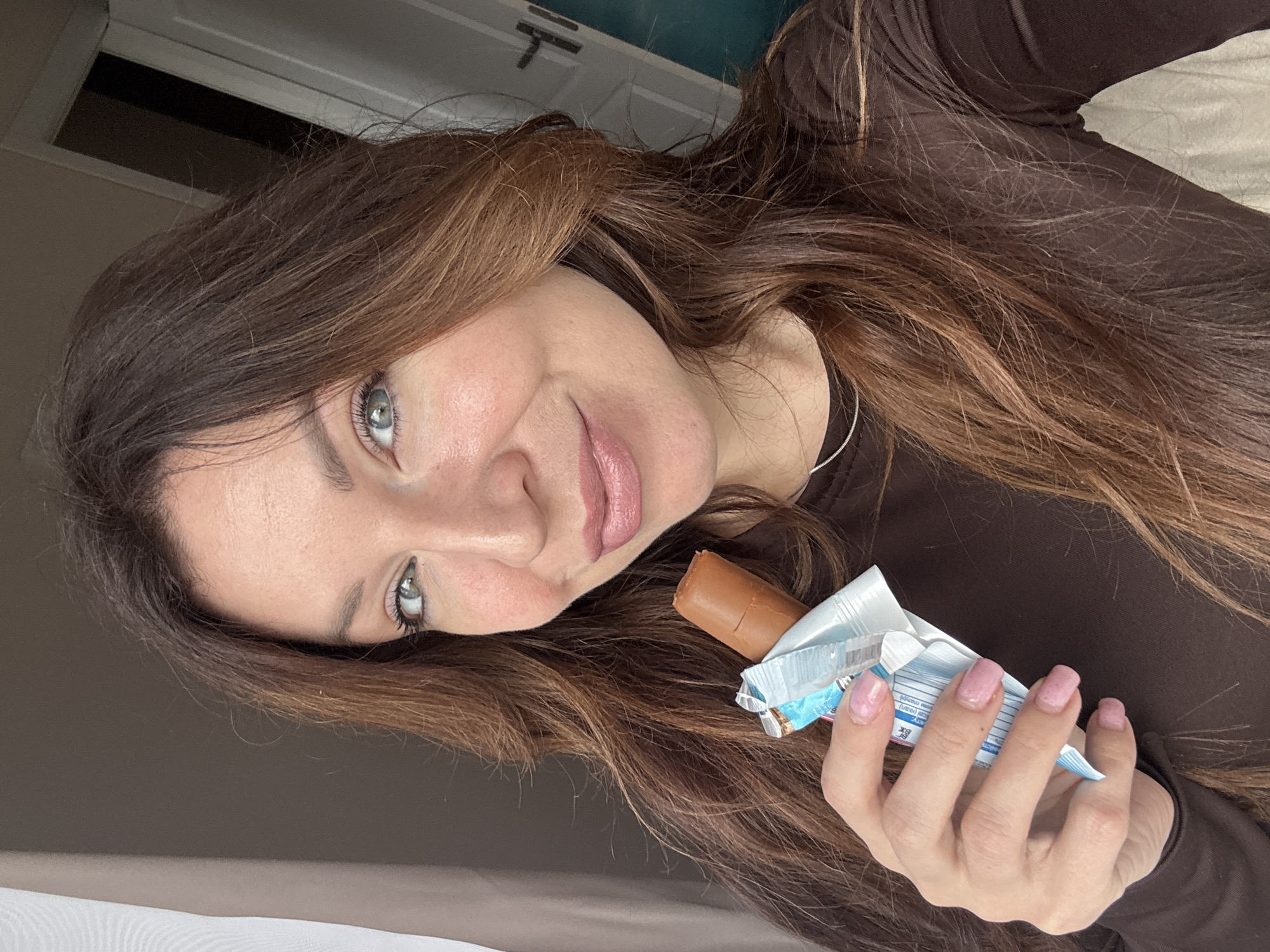 A woman with long, wavy brown hair and gray-green eyes holds a partially unwrapped chocolate bar. She is wearing a brown long-sleeved shirt and a delicate silver necklace. Her fingernails are painted a light pink color. The background includes a partially visible white door and a beige surface. The location is not specified in the image.
