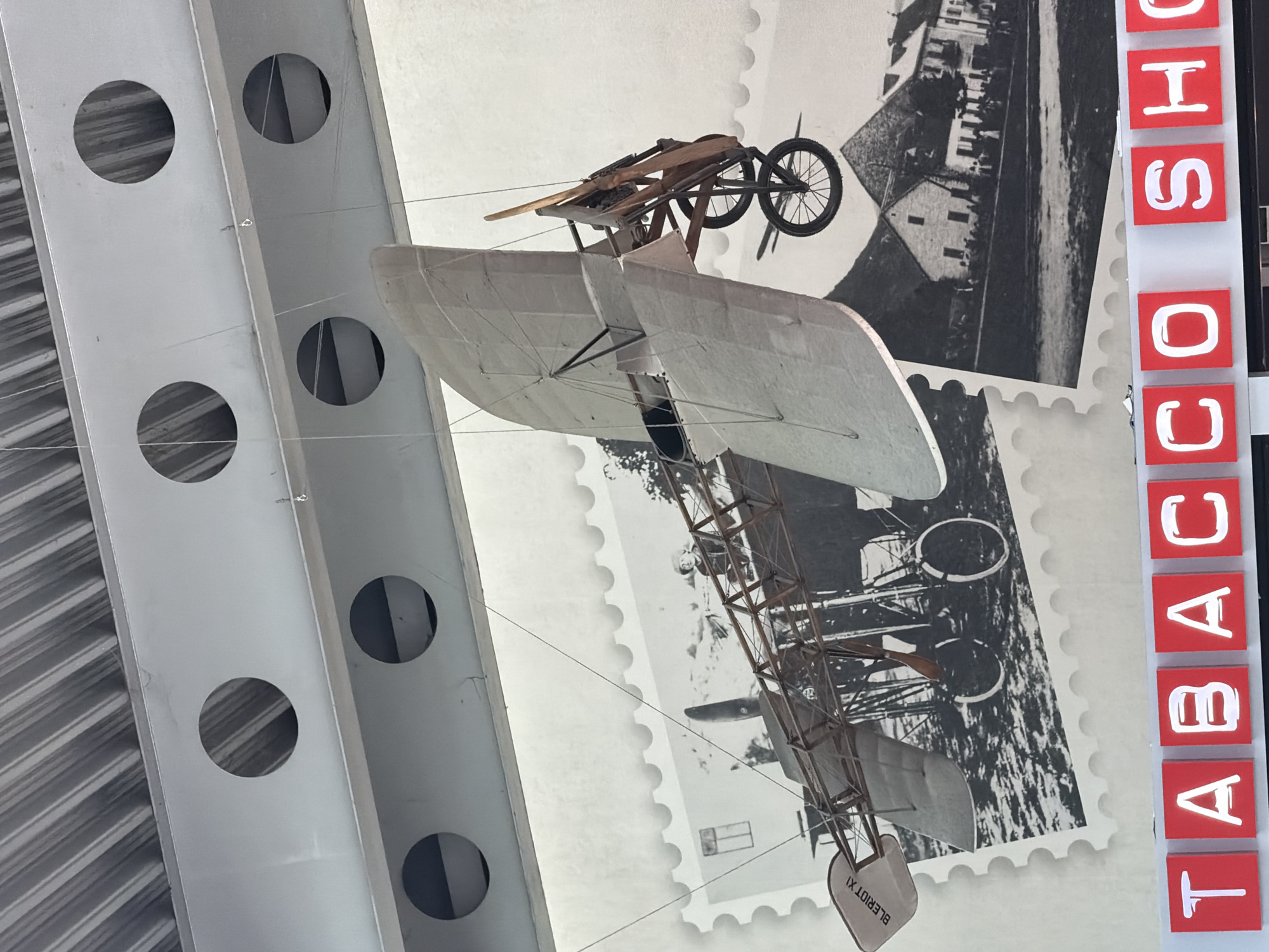 The image depicts a suspended model of an early biplane, likely a replica of the Blériot XI, hanging from the ceiling of an indoor space. The biplane is constructed with a wooden frame and fabric wings. It is positioned in front of a mural or large print that resembles a giant postage stamp, featuring historical black and white images. One image shows a crowd of people and buildings, and another shows what appears to be a field with a rudimentary structure and possibly a person. The biplane model has the text "BLERIOT XI" visible on its tail. Below the mural, red tiles with white letters spelling out "TABACCO SHO" are visible, suggesting the location is a tobacco shop. The background also shows a corrugated metal ceiling and structural beams with circular cutouts. The overall impression is a display within a commercial establishment, possibly a vintage-themed shop. The image does not provide any specific visual cues to pinpoint the exact location within the city of Donja Cijevna, Montenegro, beyond the implied nature of the establishment.