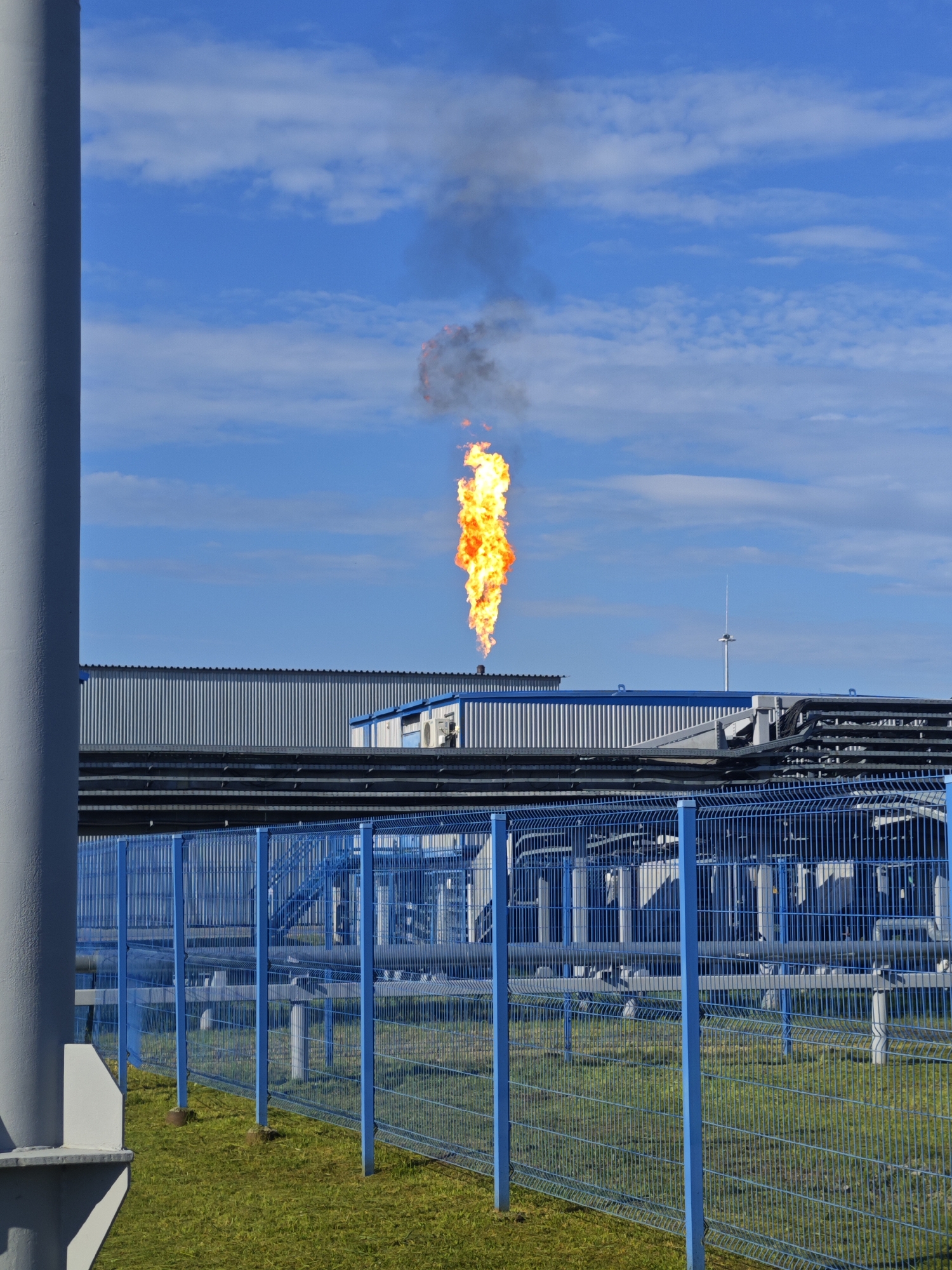 An outdoor, eye-level, medium shot captures an industrial scene under a blue sky with scattered clouds. In the center of the frame, a large flame erupts from an industrial stack, emitting a plume of dark smoke. In the foreground, a bright blue metal fence with vertical slats encloses a grassy area. Beyond the fence, industrial buildings with corrugated metal siding and blue trim are visible, along with a network of pipes and conduits. A tall, grey cylindrical structure stands on the left side of the image, partially obscuring the view of the industrial buildings. The overall impression is one of an active industrial facility.