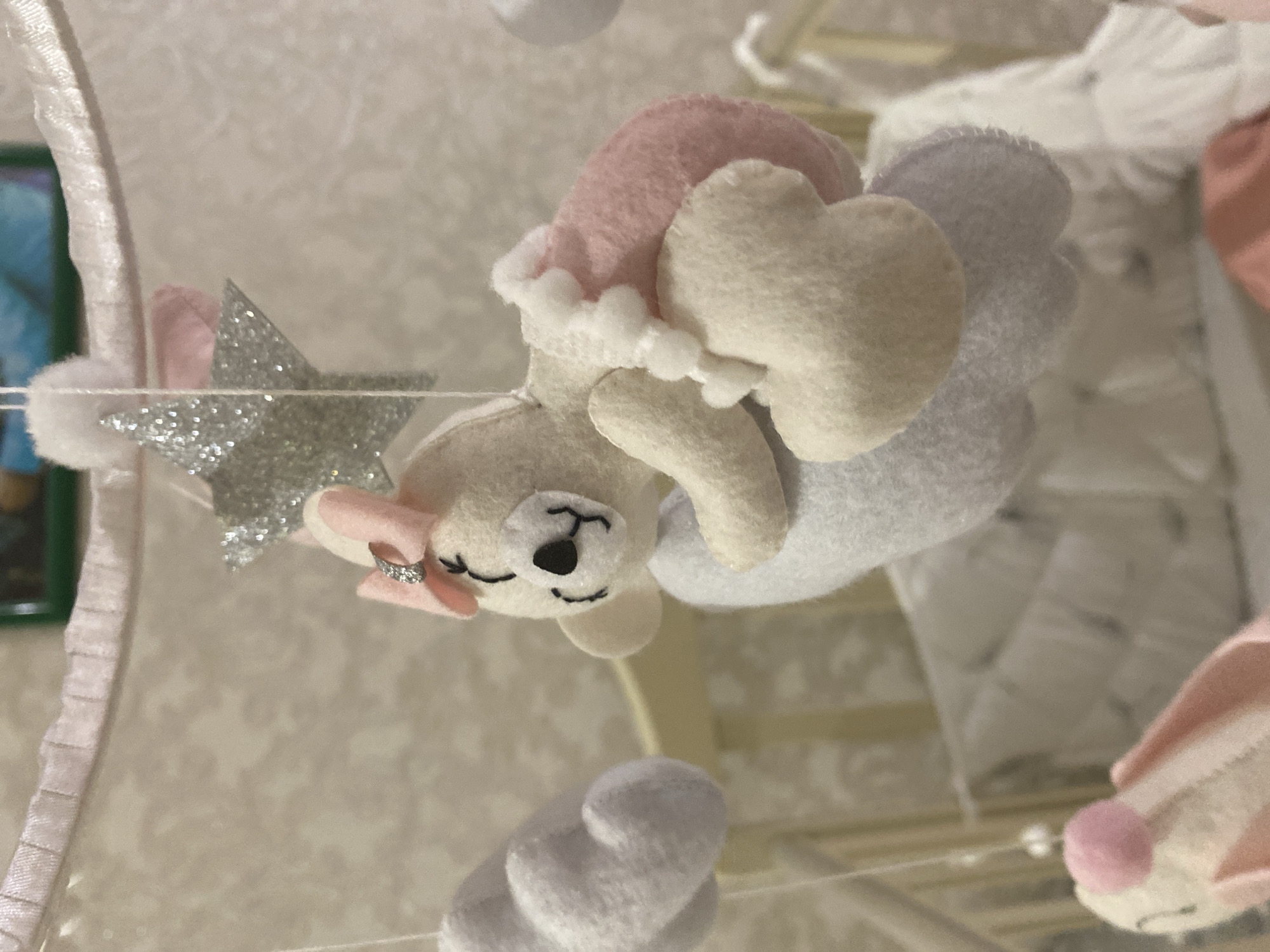The image displays a close-up perspective of a baby mobile, likely situated above a crib indoors. The central element is a cream-colored felt animal, possibly a bear, resting on its back on a light gray felt cloud. The animal features simple black embroidered closed eyes, a black nose, and a slight stitched smile. A small pink bow with a silver, possibly rhinestoned, accent is attached to its head. Its lower body is covered by a pink felt component adorned with a vertical line of small white pom-poms or bobbles.
To the left of the main figure, a white felt cloud is partially visible. Above the central figure, a silver glitter-covered star hangs, topped by a small white pom-pom. All hanging elements are suspended by thin white strings from a white-wrapped circular frame with a ruffled light pink trim along its inner edge, positioned at the top of the mobile structure. A portion of another felt character, appearing to be a bunny with pink ears and a pink pom-pom, is visible in the lower-left foreground.
The background reveals blurred light-colored wooden or plastic crib rails. Inside the crib, white bedding with a faint pattern is discernible. The wall behind the crib is a light, possibly beige, color with a subtle, repeating textured or patterned wallpaper design. A small, indistinct segment of a green-framed object and a blue hue is visible at the upper edge of the background.
