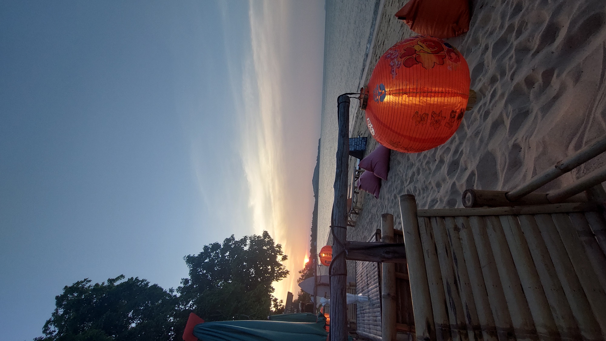The image depicts a sunset at the beach in Baan Mae Nam, Thailand. An orange paper lantern is prominently displayed. The bamboo furniture and a beach umbrella are also visible. The sky is a blend of blue and orange.