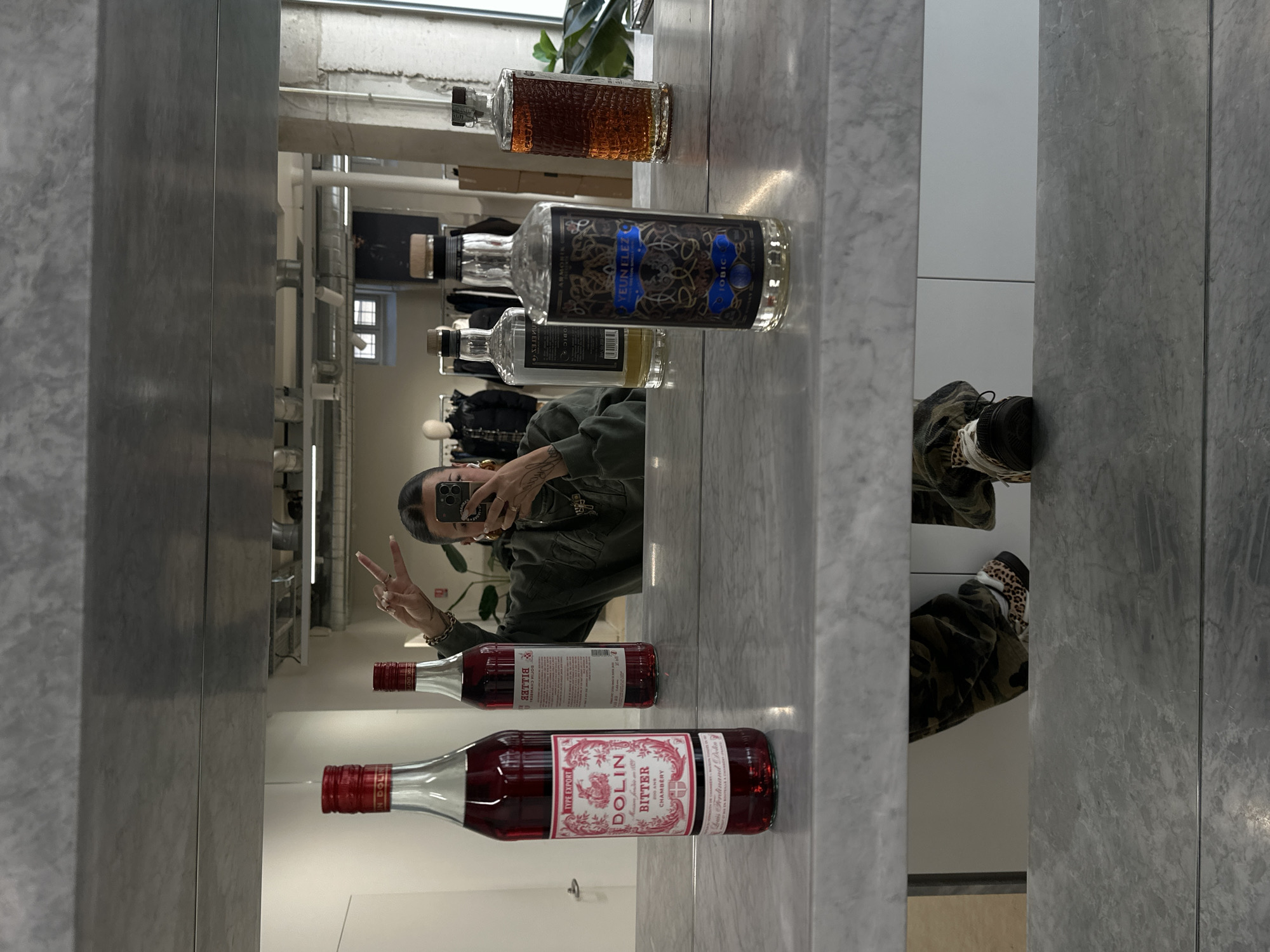 A person is visible in the mirror on a shelf with several bottles. They are taking a selfie, with a peace sign gesture. The bottles consist of Dolin Bitter Chambery, Yelin Fleet Tobie, and other various bottles of liquors. The room appears to be a clothing store with clothing racks in the background. It can be assumed that this photo was taken in Paris, France.
