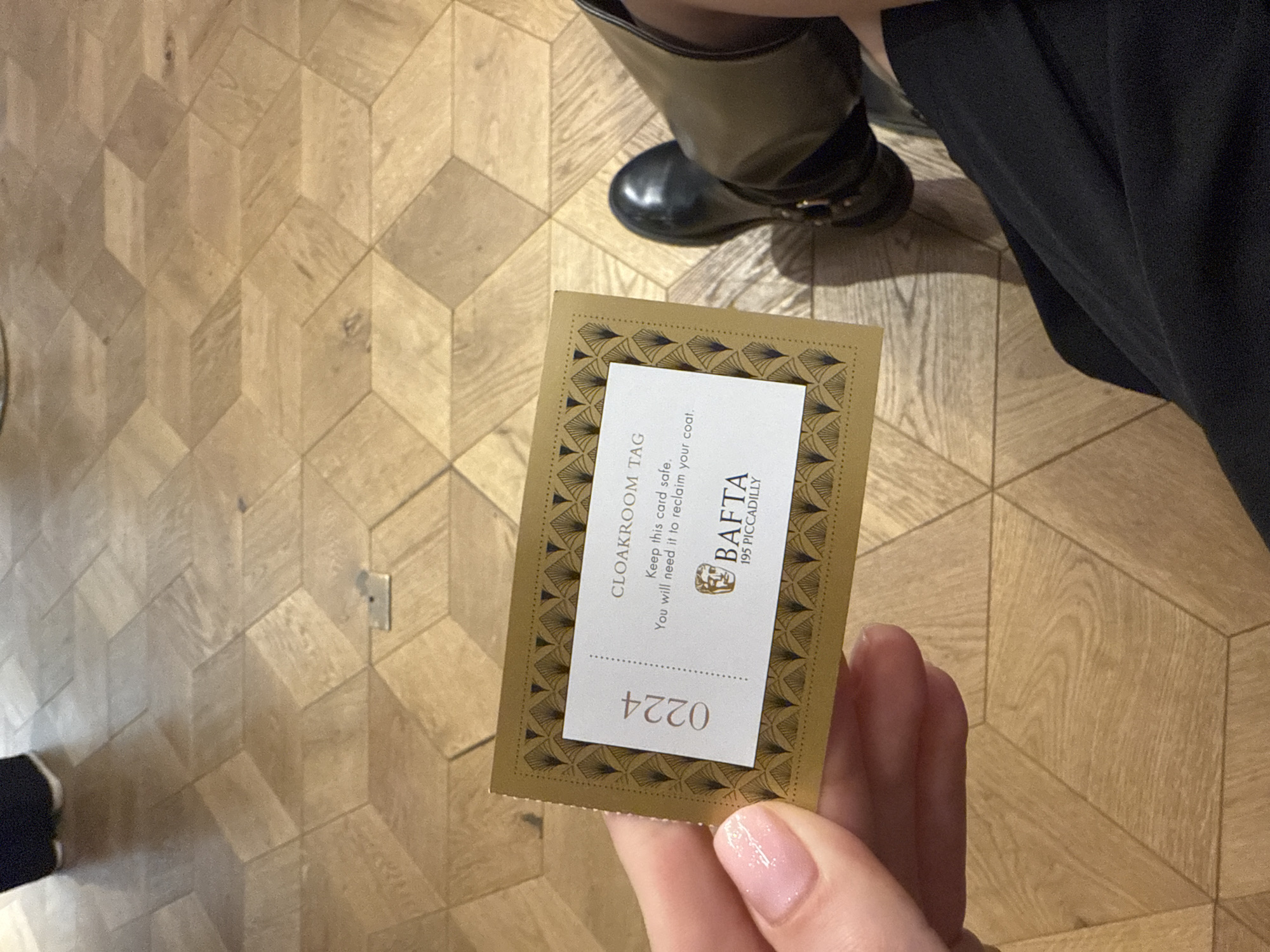 An eye-level shot captures a hand holding a cloakroom tag indoors, set against a wooden floor with a distinctive geometric, three-dimensional cube pattern. The hand, with neatly manicured nails, holds a rectangular tag featuring a gold, art deco-inspired border and a white central panel.
The visible text on the tag, partially upside down, reads "0224". The right-side-up text clearly states "CLOAKROOM TAG" and provides instructions: "Keep this card safe. You will need it to reclaim your coat." Crucially, the tag also identifies the location as "BAFTA 195 PICCADILLY," indicating the event is taking place at the British Academy of Film and Television Arts headquarters in London.
In the background, a person's lower leg and foot, wearing a dark boot with a buckle, are visible, suggesting another individual is standing nearby. A portion of dark clothing, possibly a skirt or trousers, is also seen. The lighting appears to be artificial, typical of an indoor venue. The image captures a moment related to a coat check service, likely at an event or gathering at BAFTA.