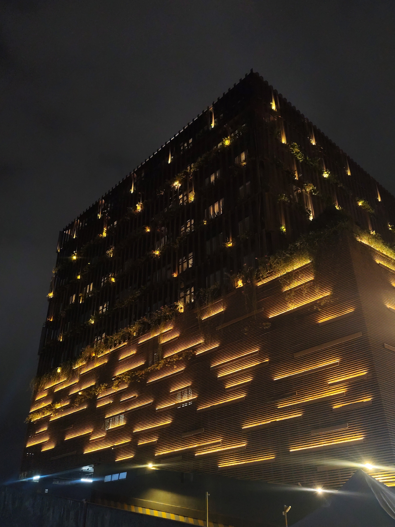 The image shows a tall, modern building at night, possibly located in Da Lat, Vietnam, based on the architectural style and available information. The building has a rectangular shape and is viewed from a low angle, emphasizing its height. Its facade features a distinctive pattern of horizontal lines, with integrated lighting that creates a visually striking effect. There are plants or greenery incorporated into the building's design, adding a touch of nature to the urban landscape. The lighting on the building is warm and golden, contrasting with the dark sky. Some light fixtures appear to be directional, casting light upwards. The foreground shows a wall or structure, and there are faint hints of street infrastructure, like a lamp post, adding to the urban environment. The sky is dark and overcast, contributing to the overall nighttime ambiance.
