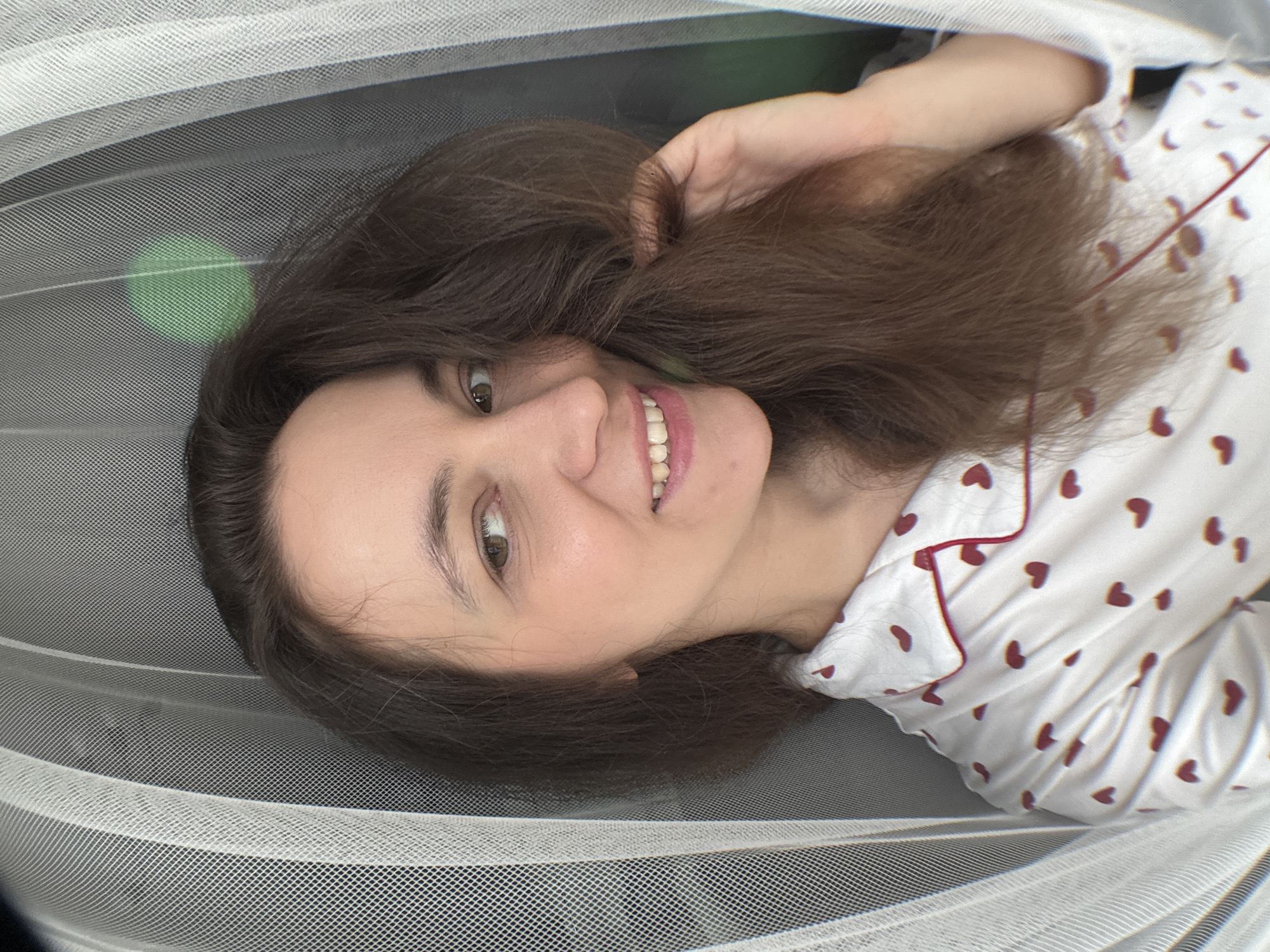 A woman with long brown hair is lying on a gray surface covered with white netting. She is wearing a white top with small red hearts and red trim. She is smiling and looking at the camera. The location is Chaikovsky, Russia.