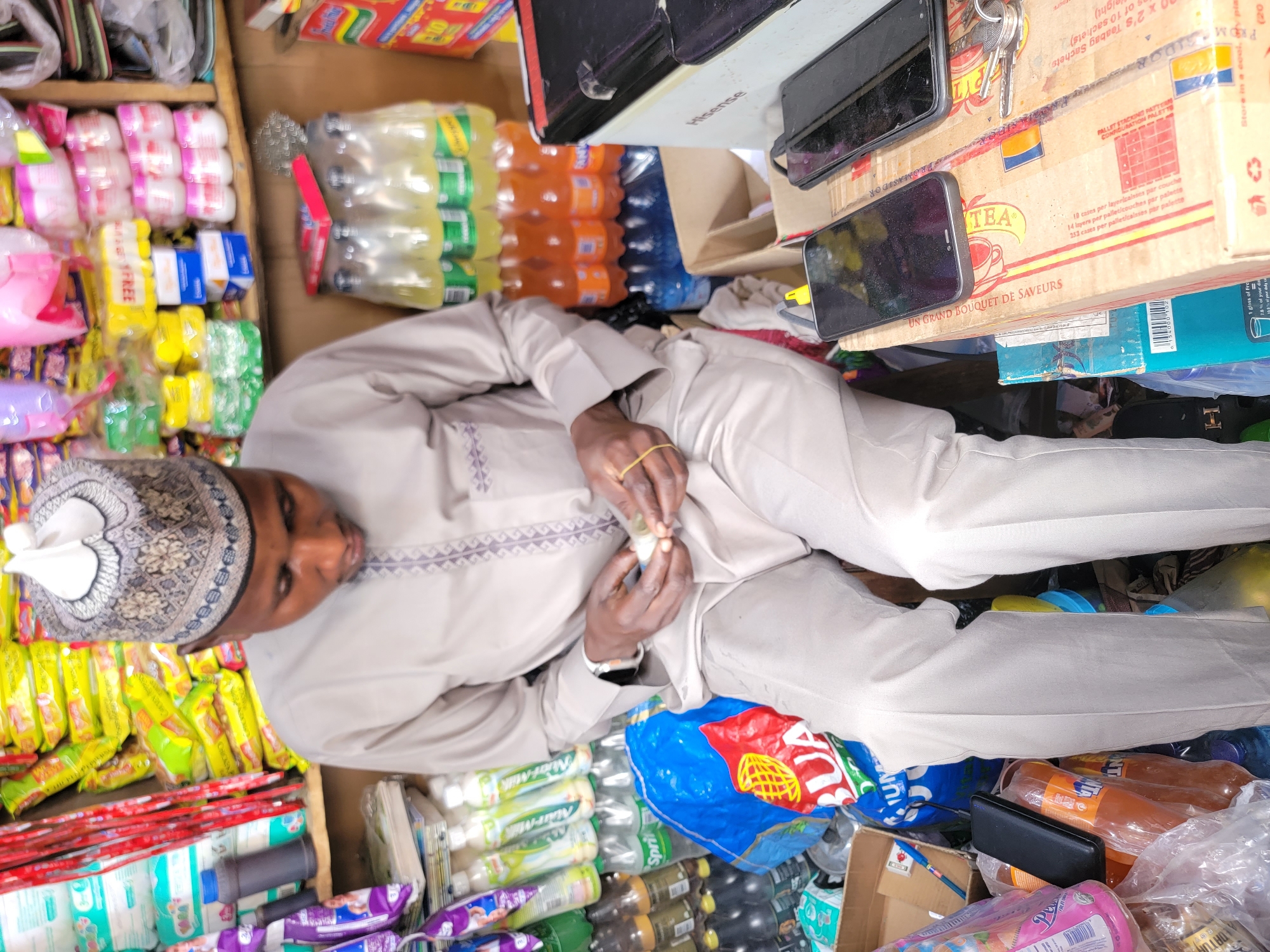 The image shows a man, presumably in Maiburiji, Nigeria, wearing a traditional cap and a light-colored garment, likely a kaftan. He appears to be handling small, rolled items. The scene is densely packed with various goods, suggesting a shop or market stall. There are numerous packaged food items, including instant noodles, bottled drinks (Sprite, Fanta, possibly yogurt drinks), and various snacks. Boxes and cartons are stacked around him, one displaying "TEA" and pallet stacking information. Two mobile phones are placed on top of the boxes. The environment is cramped, with goods tightly packed, suggesting a bustling commercial area. A blue plastic bag with "BUA" is visible.