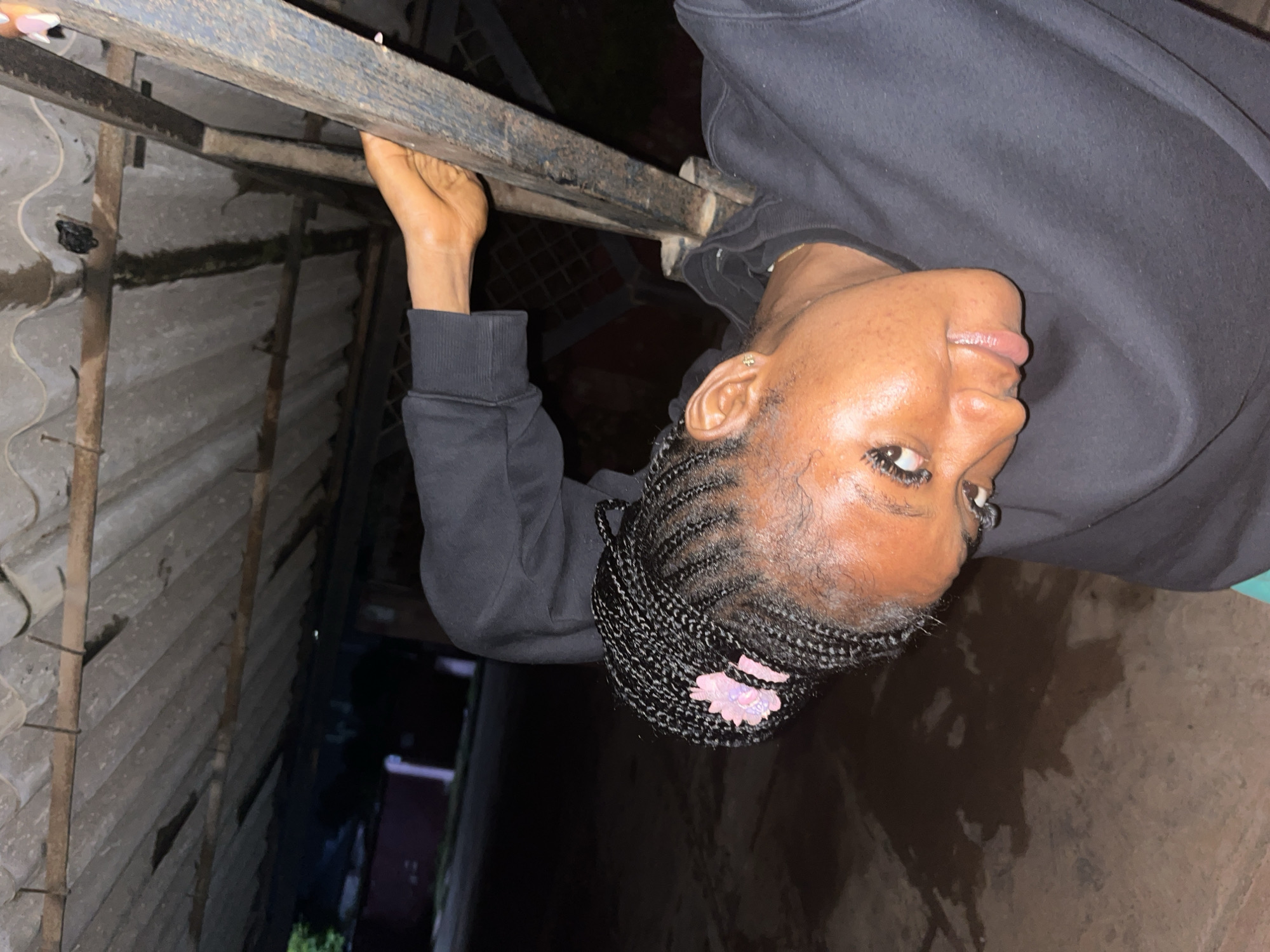 A young Black woman with braided hair adorned with a pink flower clip, wearing a black sweatshirt, is leaning against a metal support structure under a corrugated metal roof. The roof appears weathered and possibly damaged in places, with some water stains visible. The ground is uneven and appears to be wet or damp. The background suggests an outdoor urban environment, with some out-of-focus structures and vegetation visible. The lighting indicates it is nighttime or very low light conditions. The woman is looking towards the camera with her tongue slightly out. The scene is likely located in Jos, Nigeria, given the visual cues and context.