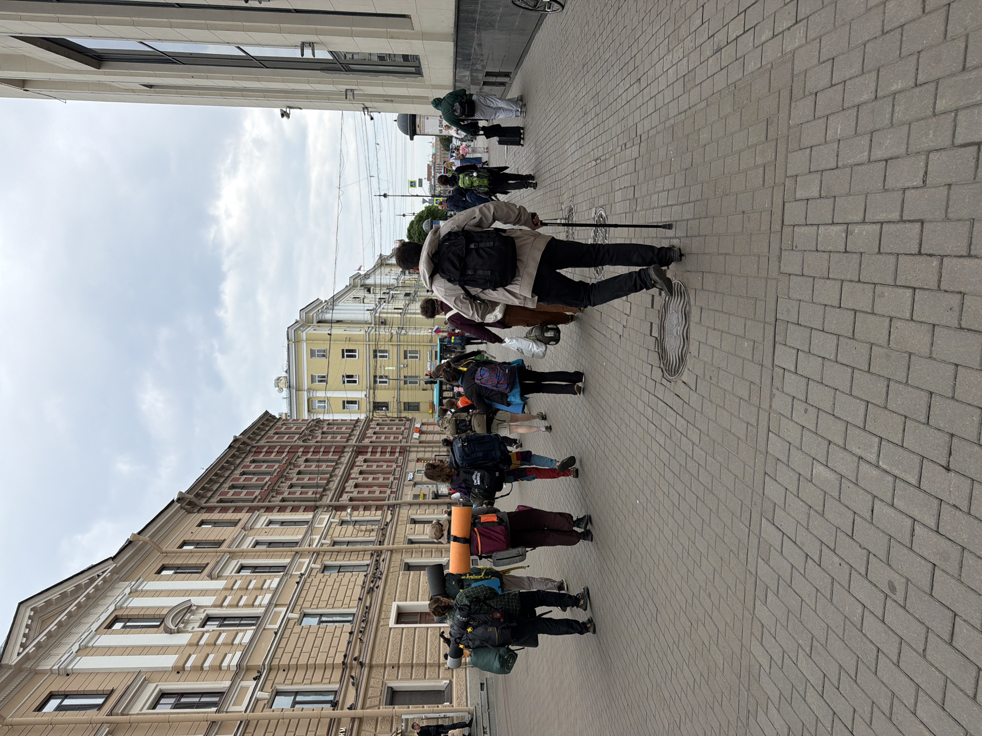 The image depicts a group of people walking along a cobblestone street in Saint Petersburg, Russia. The street is lined with multi-story buildings, one of which has a light-colored facade with architectural details and the other with a red brick facade. The sky is overcast. Many of the individuals are carrying large backpacks, suggesting they are travelers or hikers. One person in the foreground is walking with a cane. The scene appears to be a public walkway with some urban infrastructure visible, such as utility poles and streetlights.