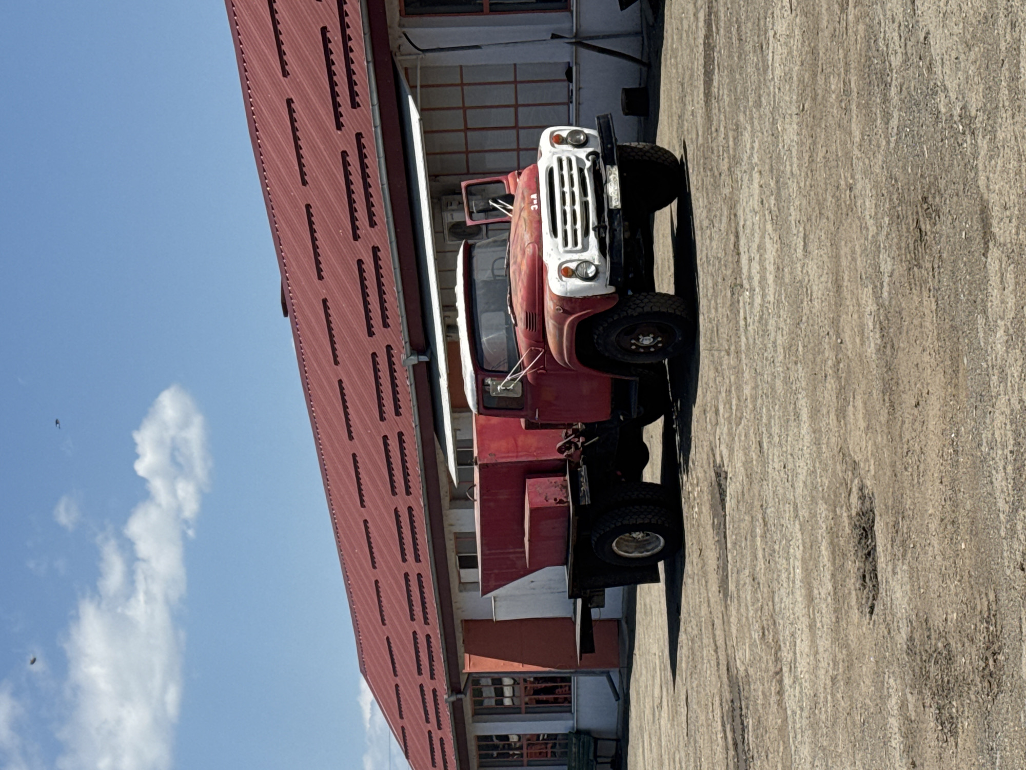 A red and white ZIL-130 truck is parked on a concrete surface next to a long, red-roofed building in Sarnevo, Bulgaria. The sky is blue with scattered clouds.
