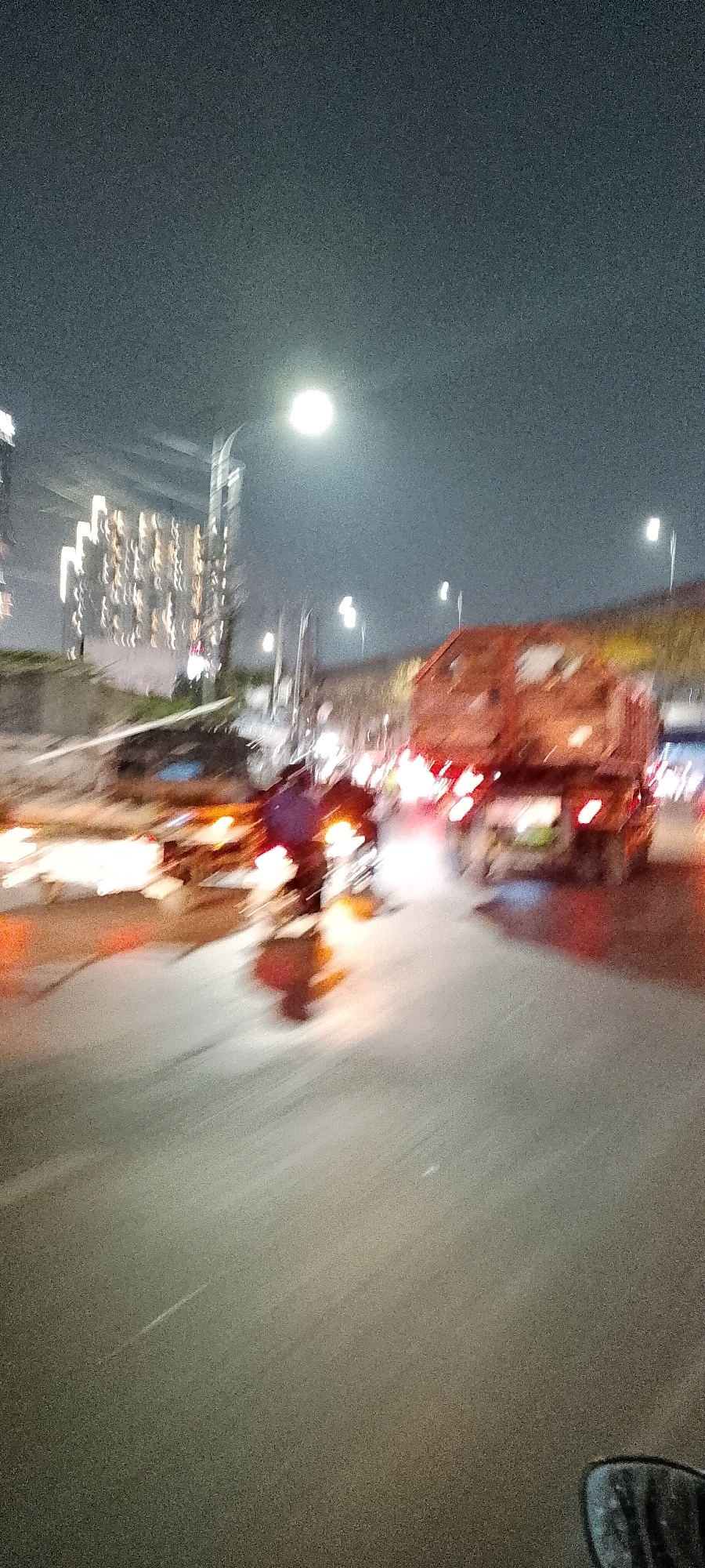 The image depicts a blurred night scene on a street in Bekasi, Indonesia. A large orange truck with a visible cargo bed is a prominent object in the right half of the frame, appearing to be in motion. In the left half, a motorcycle with a rider wearing a dark blue shirt is also captured in motion, with its headlights illuminated. Other vehicles and streetlights are visible as streaks of light due to the motion blur. In the background, the lights of a building or complex are visible, suggesting an urban environment. The sky is dark with a bright, circular light, possibly the moon or a streetlight, in the upper center of the image. The overall impression is that of a busy street at night, with vehicles moving at speed, causing significant motion blur.