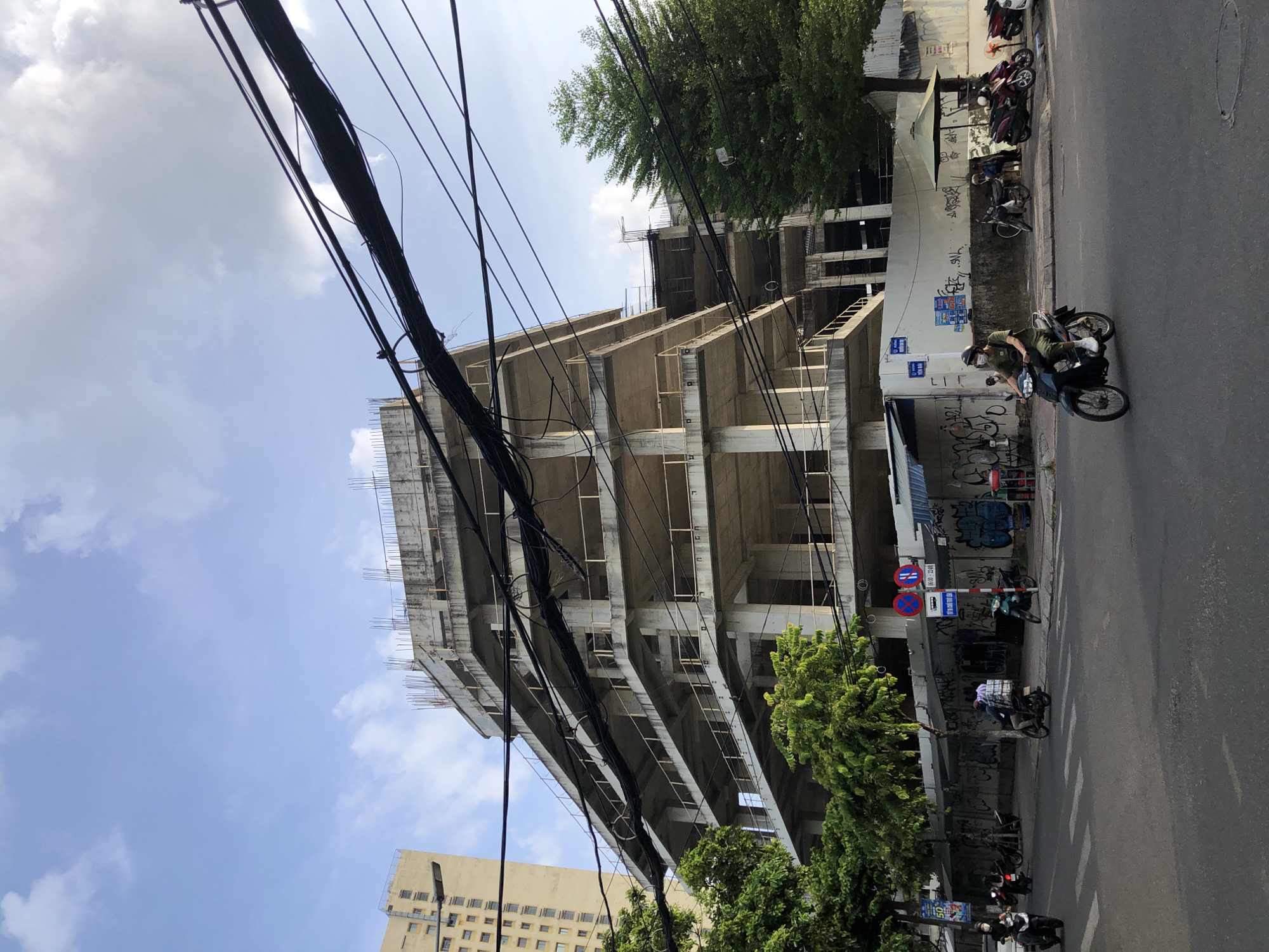 The image shows a partially constructed concrete building in Đà Nẵng, Vietnam. The building has multiple floors with exposed rebar at the top, suggesting it is still under construction. Overhead power lines are visible in the foreground, crossing the frame diagonally. On the street below, several motorbikes are present, some with riders. A few pedestrians are also visible on the sidewalk. The street is paved, and there are trees and a building in the background. The sky is blue with some clouds. The overall scene depicts an urban environment with ongoing construction and daily life.