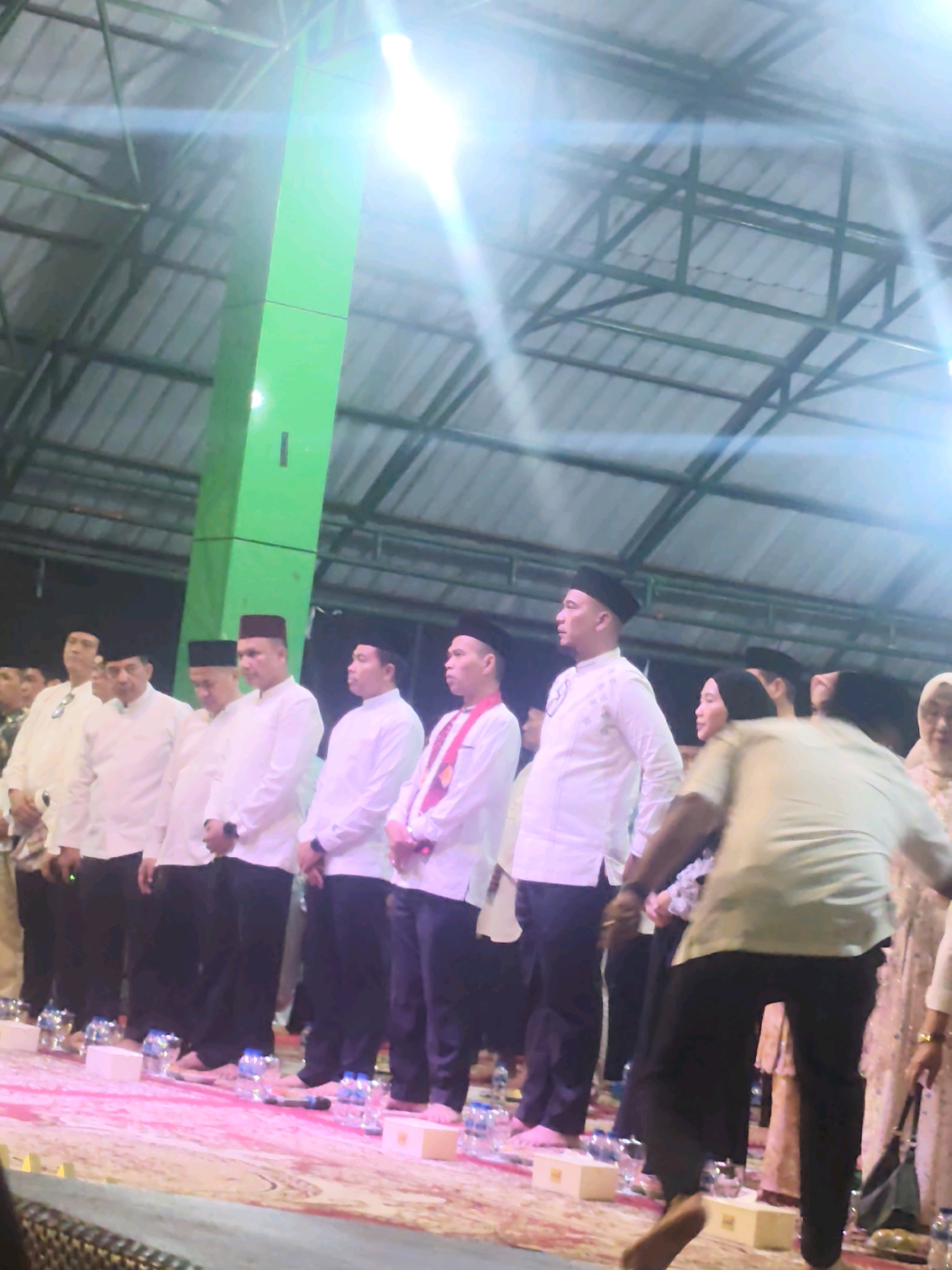 A group of men in white shirts and dark pants, some wearing traditional peci hats, stand in a line on a pink carpeted stage. They appear to be at an event, possibly a formal gathering or ceremony. To the left, a tall, bright green structure, possibly a pillar or part of a stage decoration, is visible. The background shows a dark ceiling with exposed metal beams. Bright lights are illuminating the scene from above, creating lens flares. Bottles of water and small boxes are placed on the stage in front of the men. One person in the foreground is walking away from the camera, their back to the viewer, wearing a light-colored shirt and dark pants. The overall impression is of an indoor event held at night, likely in a covered outdoor or semi-outdoor venue in Rengat, Indonesia, given the context.