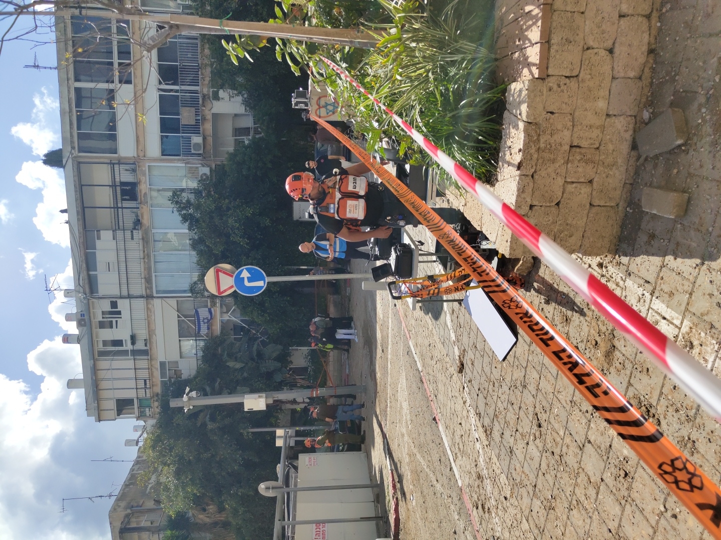 The scene depicts an outdoor emergency response operation during daylight hours on a paved street in an urban setting, identified as Tel-Aviv, Israel. The ground is covered with scattered dirt, small stones, and broken paving bricks.
The immediate foreground features red and white striped barrier tape and orange and black striped barrier tape. The orange tape displays Hebrew text "אזור סכנה! אין כניסה" (Danger Zone! No Entry) and a Star of David emblem. Behind this tape, a male individual in an orange helmet, dark shirt, and an orange and black reflective vest with "חובש" (Medic) in Hebrew is partially visible, facing towards the left. A foldable table with electronic equipment and a white rectangular panel lie on the ground within the cordoned area.
In the mid-ground, several individuals, presumed to be emergency personnel, are present. One male in a blue reflective vest is observed looking down at a device. Further back, a group of individuals stands near a light-colored portable structure with "סכנה! אין כניסה" (Danger! No Entry) written in Hebrew, and a dark sedan vehicle is partially visible. A street light pole with a CCTV camera is present, along with two circular road signs: a yield sign and a mandatory right turn sign.
The background is dominated by a multi-story residential building featuring multiple balconies with metal railings, large windows, and external air conditioning units. An Israeli flag is suspended from a second-floor balcony. Various trees and shrubs are situated throughout the scene, partially obscuring parts of the buildings and mid-ground. The sky is partially cloudy.