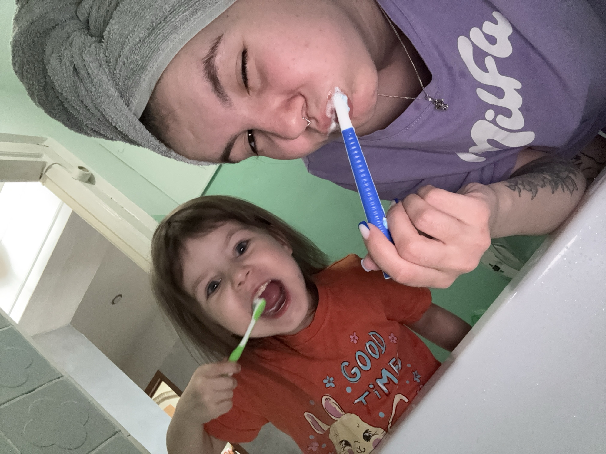 The image depicts a close-up, low-angle indoor scene in what appears to be a bathroom in the city of Фоки, Russia. Two individuals are engaged in brushing their teeth. On the right, an adult female is present, wearing a grey towel wrapped around her head and a light purple t-shirt with "Nilfa" (or similar script) printed in white. She has a silver chain necklace with a small pendant, a septum nose piercing, and a tattoo on her left forearm. She holds a blue toothbrush in her right hand, actively brushing her teeth with visible foam, and winks with her right eye. To her left, a young child, likely female, is visible, wearing an orange t-shirt featuring a cartoon rabbit and the text "GOOD TIME" in blue. The child holds a green toothbrush in her right hand, actively brushing her teeth with her mouth open and a wide smile. A white basin or counter surface is partially visible in the lower foreground with water droplets. The background consists of a light green wall and a white ceiling with textured panels in the upper left, as well as a partial view of a doorway with a brown frame. The scene is brightly lit.