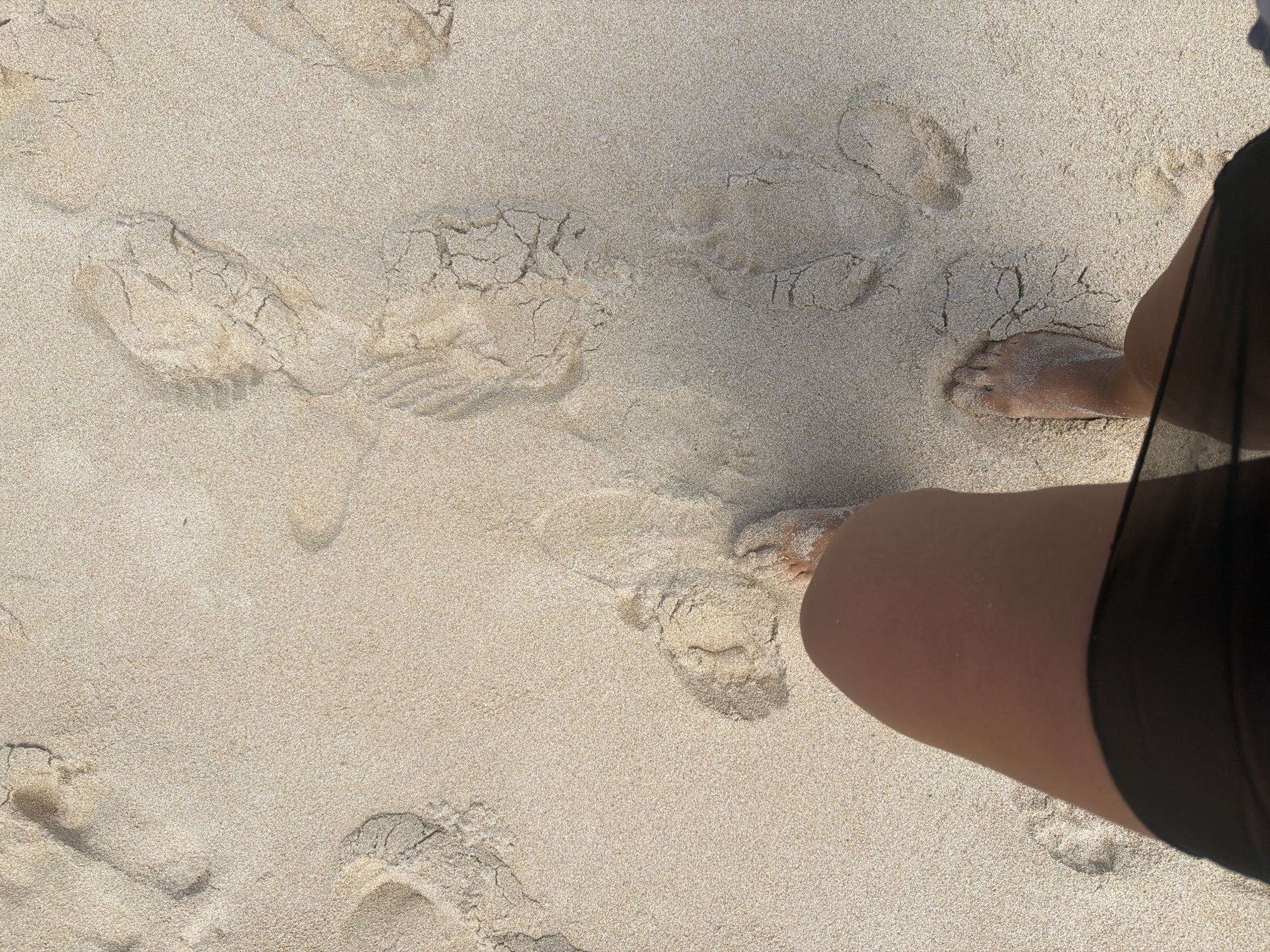The image depicts a sandy beach, likely in Ungasan, Indonesia. The scene shows various footprints in the sand, indicating recent activity. A person is visible on the right side of the frame, with their bare feet partly buried in the sand. They are wearing dark-colored attire. The sand is light in color and appears dry, with slight variations in texture due to the footprints. The lighting suggests it is daytime, possibly mid-morning or afternoon. The overall setting is a beach environment with signs of pedestrian traffic.