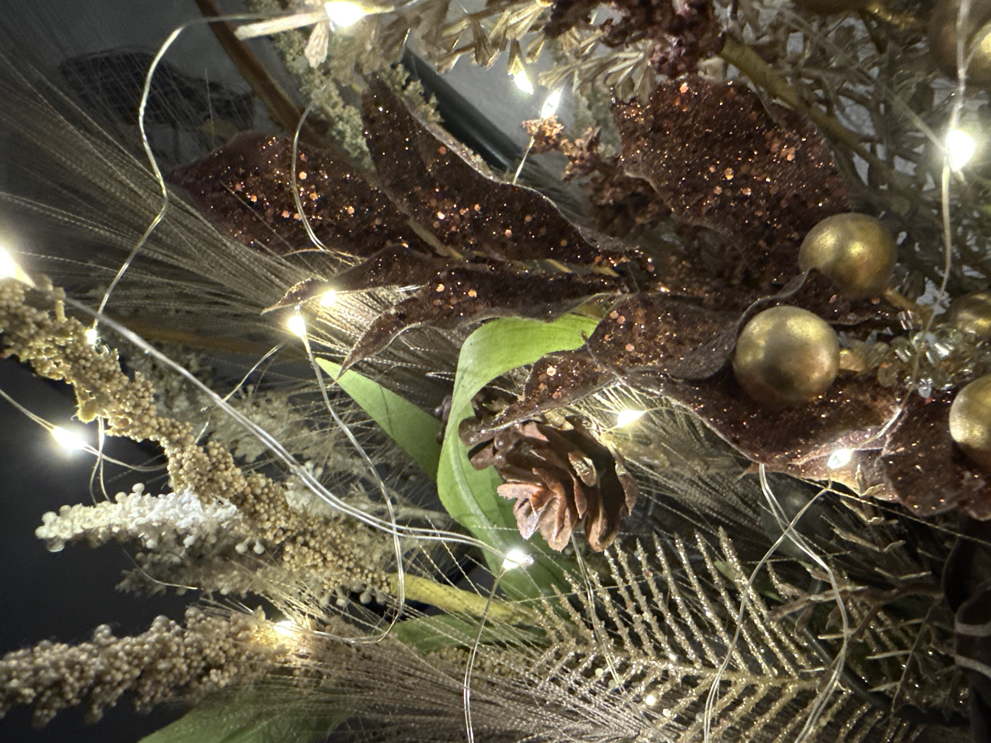 A close-up vertical shot displays a festive or decorative arrangement, likely a holiday or seasonal display, set against a dark, indistinct background. The arrangement is illuminated by numerous small, warm-toned LED string lights with thin, clear wires, casting bright highlights on various elements. Dominant colors include shades of brown, gold, beige, and muted green.
Key objects observed are:
- **Dried Floral/Plant Material:** Several upright stalks of beige and light brown dried plant material with a fuzzy or seeded appearance are prominent in the upper-left, upper-right, and central areas. Some resemble dried grasses or plumes.
- **Glittery Foliage:** Dark brown, artificial leaf-like or petal-shaped elements heavily coated in gold or copper glitter are interspersed throughout the center and right side of the frame.
- **Pinecone:** A single small, brown pinecone is positioned slightly to the left of the image center.
- **Green Leaves:** Two distinct, broad, vibrant green leaves are visible; one to the left of the pinecone and another partially visible in the upper-center.
- **Metallic Balls:** Several round, matte gold decorative balls are present, primarily in the lower-left and lower-right quadrants.
- **Metallic Fern:** A flattened, metallic gold, fern-shaped decorative piece is visible in the lower-left portion of the frame.
- **String Lights:** The small LED lights are randomly distributed, with their thin wires creating curvilinear lines across the composition.
The scene depicts a static arrangement, with no discernible human presence or active interactions. The environment appears to be an interior setting, possibly part of a larger decorative display.
Location: Unknown, Unknown